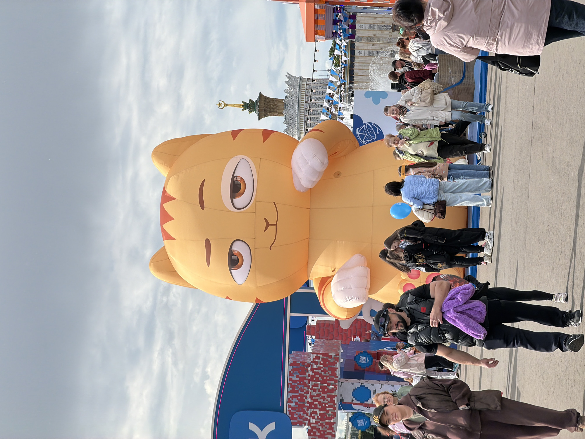 The image captures a scene at the VDNKh (Exhibition of Achievements of National Economy) in Moscow, Russia. The focal point is a large, inflatable cartoon cat, possibly an advertisement or decoration for an event. A crowd of people surrounds the cat, with some individuals appearing to take photos or interact with the exhibit. The background includes architectural elements of the VDNKh, such as the iconic central pavilion topped with a spire and star. The sky is overcast, and the general atmosphere suggests a public gathering or festival.