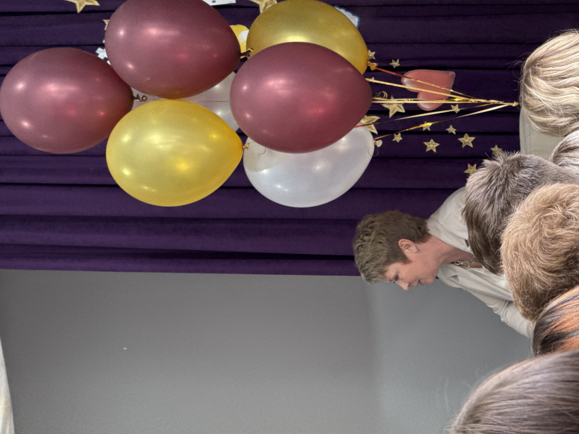 A slightly high-angle shot shows balloons, people, and decor in front of a purple draped wall over a gray wall in an interior, unspecified location. Six inflated balloons are positioned at the top left of the image, with colors including maroon, yellow, and white. Golden star-shaped decorations, along with streamers, are attached to the balloons. Several people are visible, primarily from the shoulders up, suggesting a gathering or event. A woman with short hair is shown in profile, looking down, partially obscured by other people. The wall is divided into two horizontal sections: the top portion is draped in purple fabric, while the bottom section is a solid gray. The overall setting indicates a celebratory or event-oriented environment.