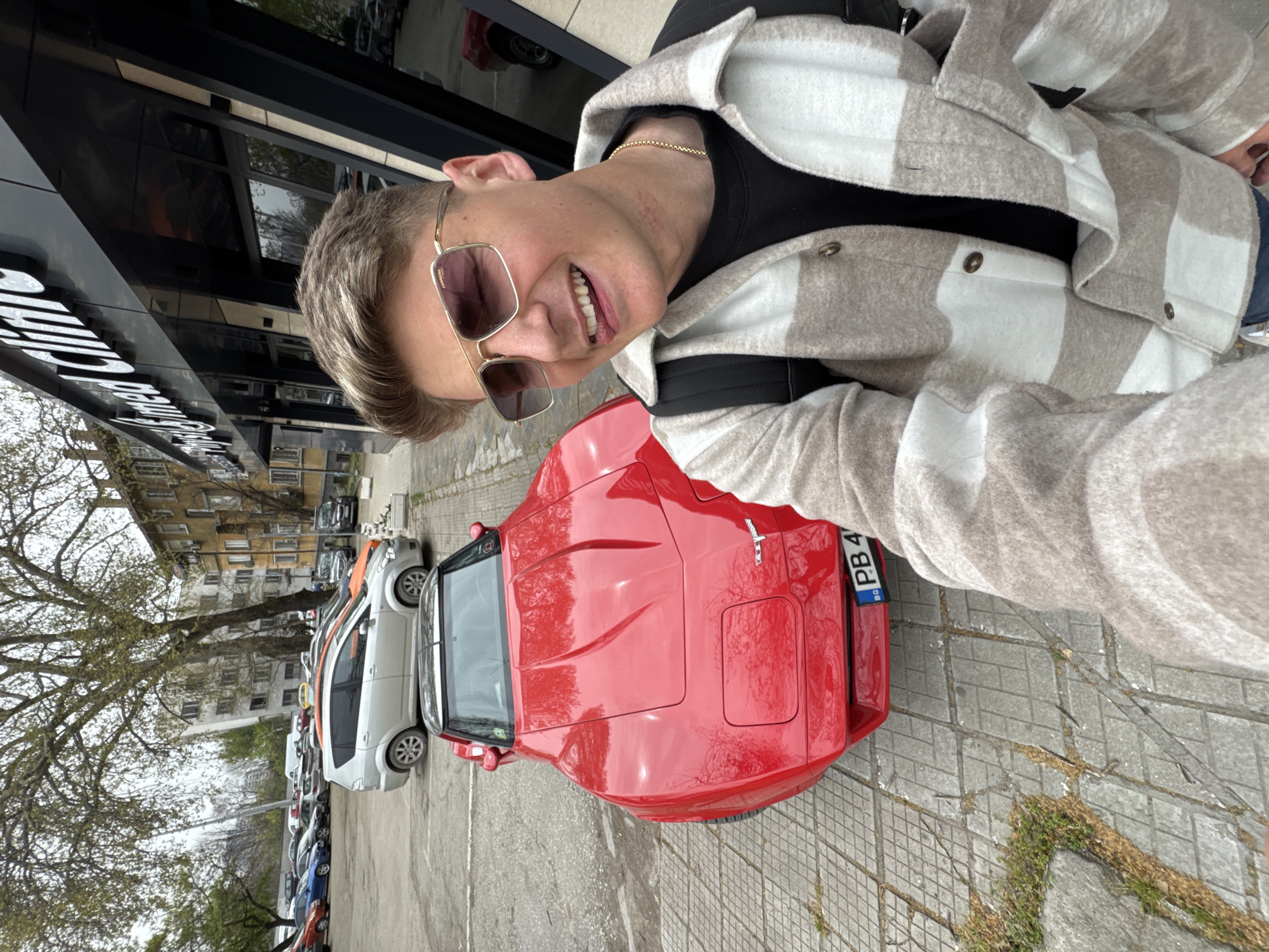 A person with short blond hair and sunglasses takes a selfie. They are wearing a black shirt under a cream and grey checked jacket and a gold chain. The subject is positioned in front of a red sports car with a Bulgarian license plate reading "PB 4." The scene appears to be on a street in Plovdiv, Bulgaria, as evidenced by the license plate. Behind the red car, a line of parked cars is visible. Trees are also present on the left side of the image. The background shows a building with multiple floors.