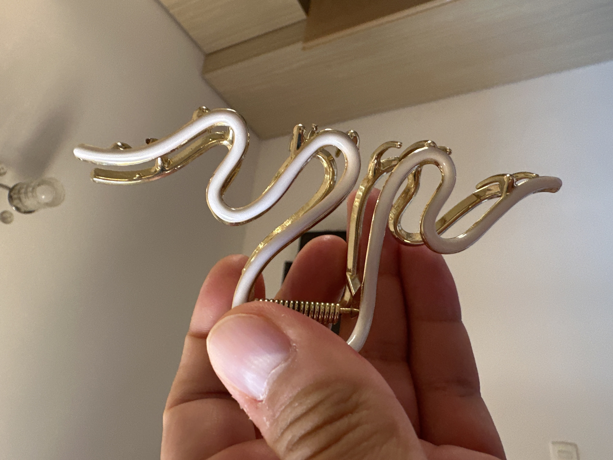 A hand is holding a gold and white snake-shaped hair clip. The background is a light-colored wall with a ceiling light fixture visible at the top. The scene appears to be indoors.