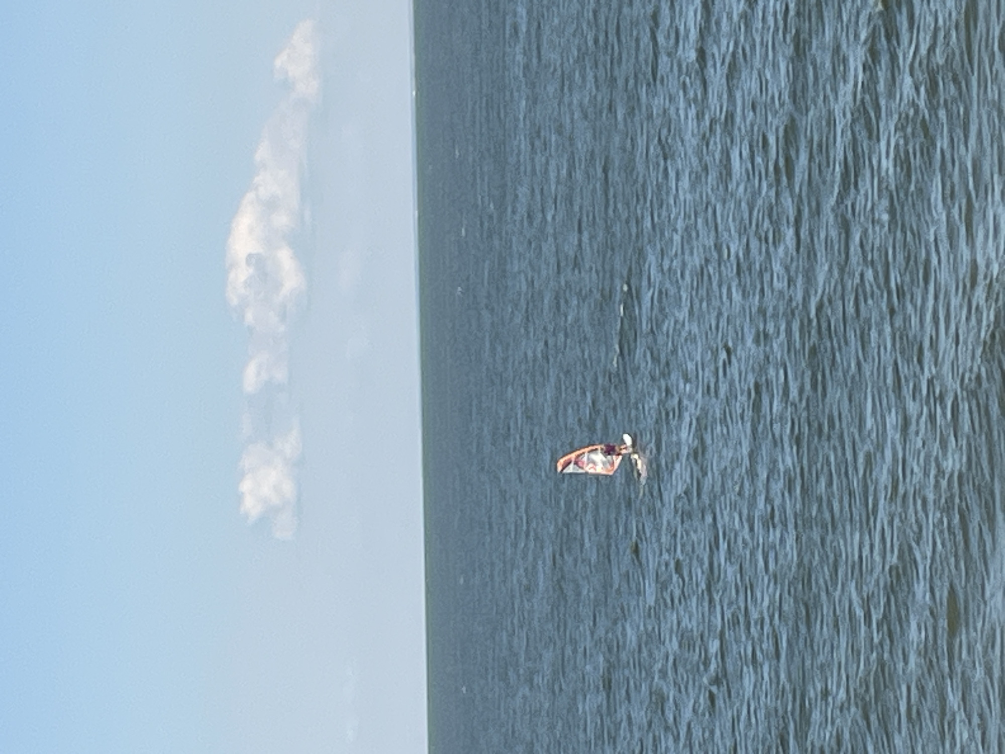 An extreme wide shot captures a single windsurfer in the middle of a vast, deep blue body of water. The windsurfer is a distant, small figure, positioned slightly off-center to the right. They are on a white board with a sail that displays a colorful, abstract design of orange, white, and dark red. The water is calm, with gentle ripples disturbing its surface, indicating a light breeze. The horizon line is sharp and distinct, separating the water from a pale blue sky that is dotted with wispy white clouds. The overall impression is one of solitude and activity in a serene, expansive natural environment. The image does not contain any elements that would allow for the determination of its specific location within Taganrog City District, Russia.