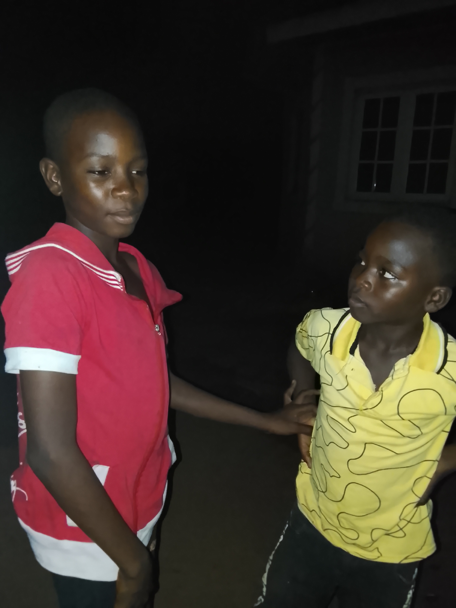 Two young boys are captured in a vertical, low-light photograph, likely taken at night, in Akwanga, Nigeria. The scene features a darker background, with a white-framed window of a building faintly visible on the right side.
The boy on the left, appearing slightly older, stands in a red polo shirt with white striped trim on the collar and white cuffs. He wears dark trousers. His face is illuminated, and he looks downwards and to his right with a contemplative expression. His right hand is gently placed on the left arm or side of the shorter boy beside him.
The shorter boy on the right wears a yellow polo shirt with black abstract, swirling patterns and a black-trimmed collar, along with dark trousers featuring white accents. He stands with his hands on his hips, gazing upwards and to his left, seemingly at the taller boy, with a look of curiosity or inquiry. Both boys have dark skin tones and closely cropped hair. The primary light source appears to be artificial, illuminating their faces and clothing against the dark surroundings. There is no visible text in the image.