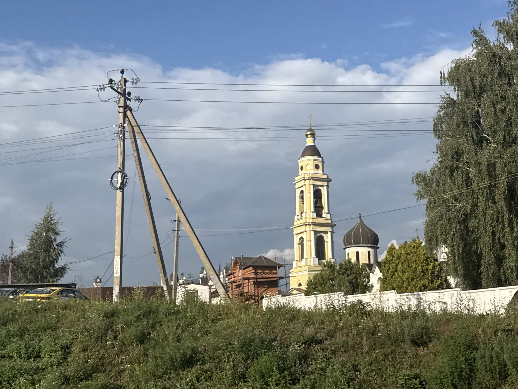In the city of Mikhailovskaya Sloboda, Russia, a serene sky with scattered clouds stretches above a religious complex. A prominent yellow bell tower with a golden cross atop a dark dome stands tall, accompanied by another dark dome belonging to a nearby building. The complex features white walls, some appearing to be under construction with scaffolding visible on a brick structure. Lush green vegetation, including tall grass and trees, blankets the foreground and surrounds the site. Power lines crisscross the scene, supported by concrete utility poles, one of which has a triangular bracing structure. A yellow car is parked in the background, partially obscured by the greenery. The overall impression is one of rural tranquility with active religious and civic infrastructure.