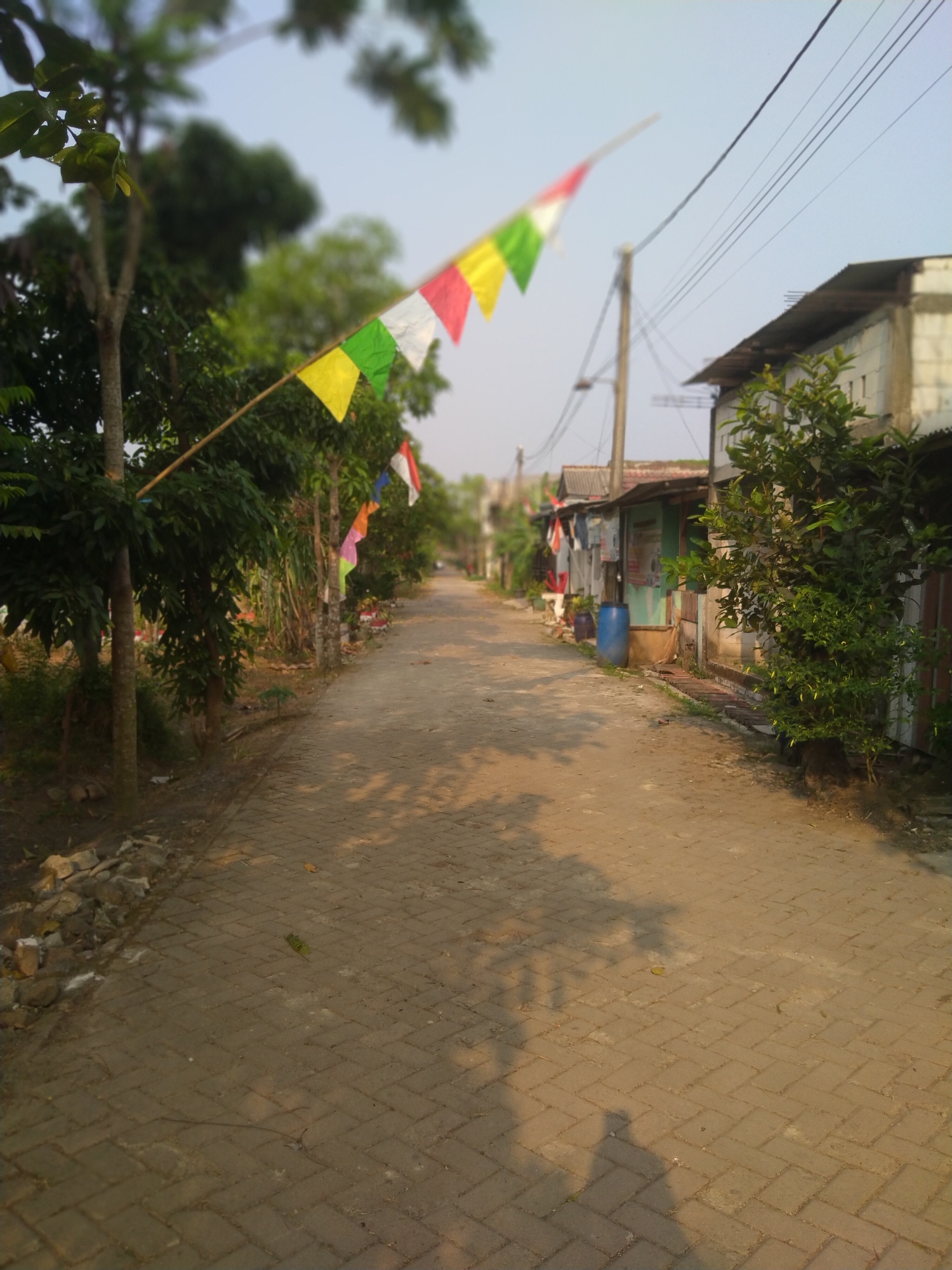 The image depicts a narrow, paved street in Cisoka, Indonesia, bordered by trees and small residential buildings. Several colorful triangular flags are strung across the street, casting shadows on the ground. A large blue barrel stands near one of the buildings, and electrical wires are visible overhead, supported by a utility pole. The scene appears to be captured during the late afternoon or early evening, as evidenced by the warm sunlight and long shadows. The area seems to be a modest, rural or semi-urban neighborhood.