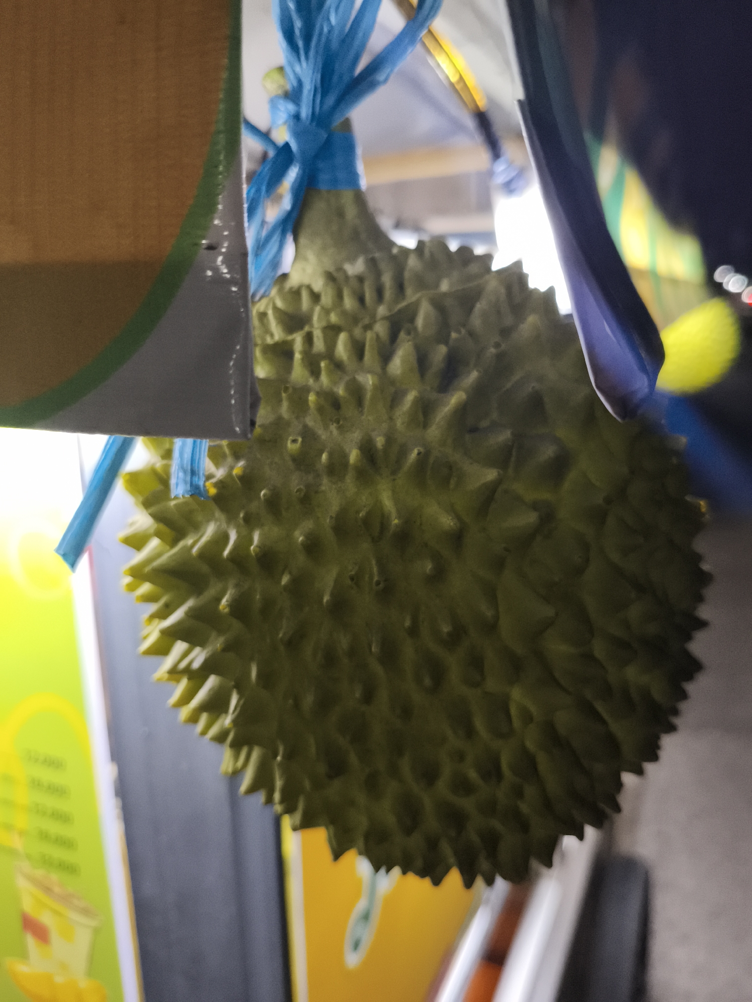 A close-up, shallow depth of field shot shows a durian fruit hanging from blue plastic ties. The fruit, a common sight in Southeast Asia, is large, spiky, and green. The background is blurred, suggesting an outdoor market setting in Depok, Indonesia, likely at night, given the artificial lighting and bokeh effects in the background. A portion of a wooden structure with a green and grey trim is visible on the left side of the frame. The durian appears to be part of a display for a street food vendor, with parts of colorful signs visible in the blurred background.