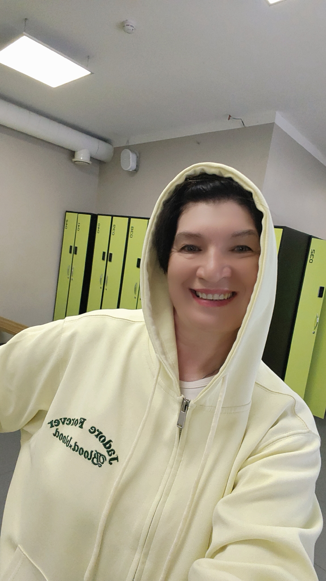 A smiling woman wearing a yellow hoodie with the hood up is posing for a selfie in what appears to be a locker room. Behind her, there are lockers painted in shades of green and yellow. A pipe and a speaker are visible on the wall near the ceiling. A rectangular light fixture is attached to the ceiling. The city of Krasnoyarsk, Russia, is not discernible from the image alone.