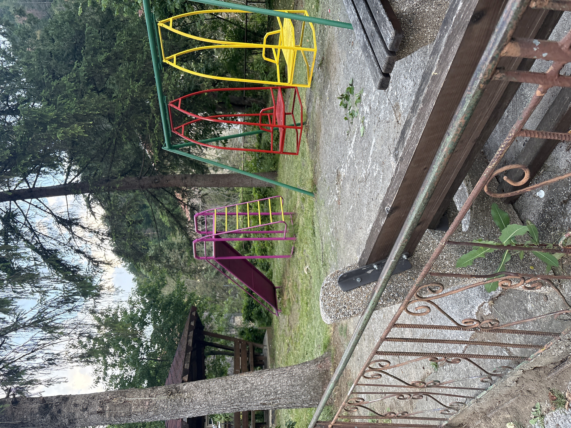 This image depicts a deserted outdoor children's playground located in Unknown, Unknown. The scene is observed from behind a rusted, ornate wrought-iron fence that spans the lower foreground, featuring decorative scrollwork and vertical bars. A weathered wooden bench with concrete supports is positioned directly behind the fence, with a horizontal green metal bar running parallel to it.
To the left, a large tree with thick, textured bark stands prominently. Behind this tree, a wooden gazebo with a dark, possibly shingled roof and internal seating is partially visible. Centrally, a purple children's slide with a yellow-railed ladder is erected on a grassy patch. To the right of the slide, a green-framed swing set features two swings: one red and one yellow, both designed with elevated backs and side supports. The ground beneath the swings and around the slide consists of a patchy mix of concrete or compacted dirt and sparse grass, with some fallen leaves and debris.
The background is dominated by dense green foliage and numerous trees, suggesting a natural or wooded area. A cloudy sky is faintly visible through the tree canopy at the top. No individuals are present in the scene. The playground equipment, fence, and bench exhibit signs of age and wear, including rust on the metal components and weathered surfaces on the wood.