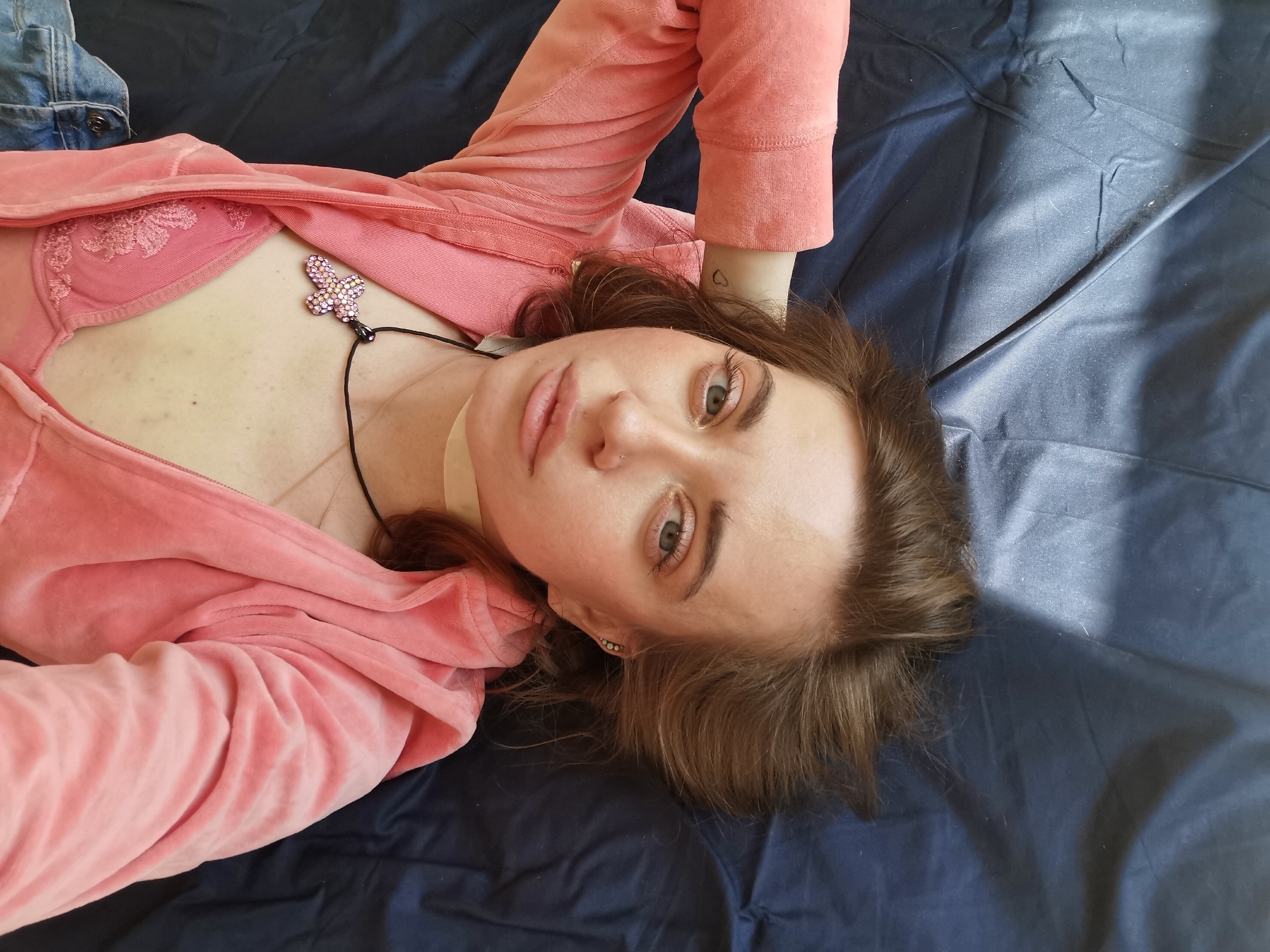 A young woman lies on a dark blue sheet. She is wearing a pink velour tracksuit and a pink bra. She is wearing a necklace with a butterfly pendant. Her hair is brown and shoulder-length. She has a small heart tattoo on her wrist. The location is not discernable.