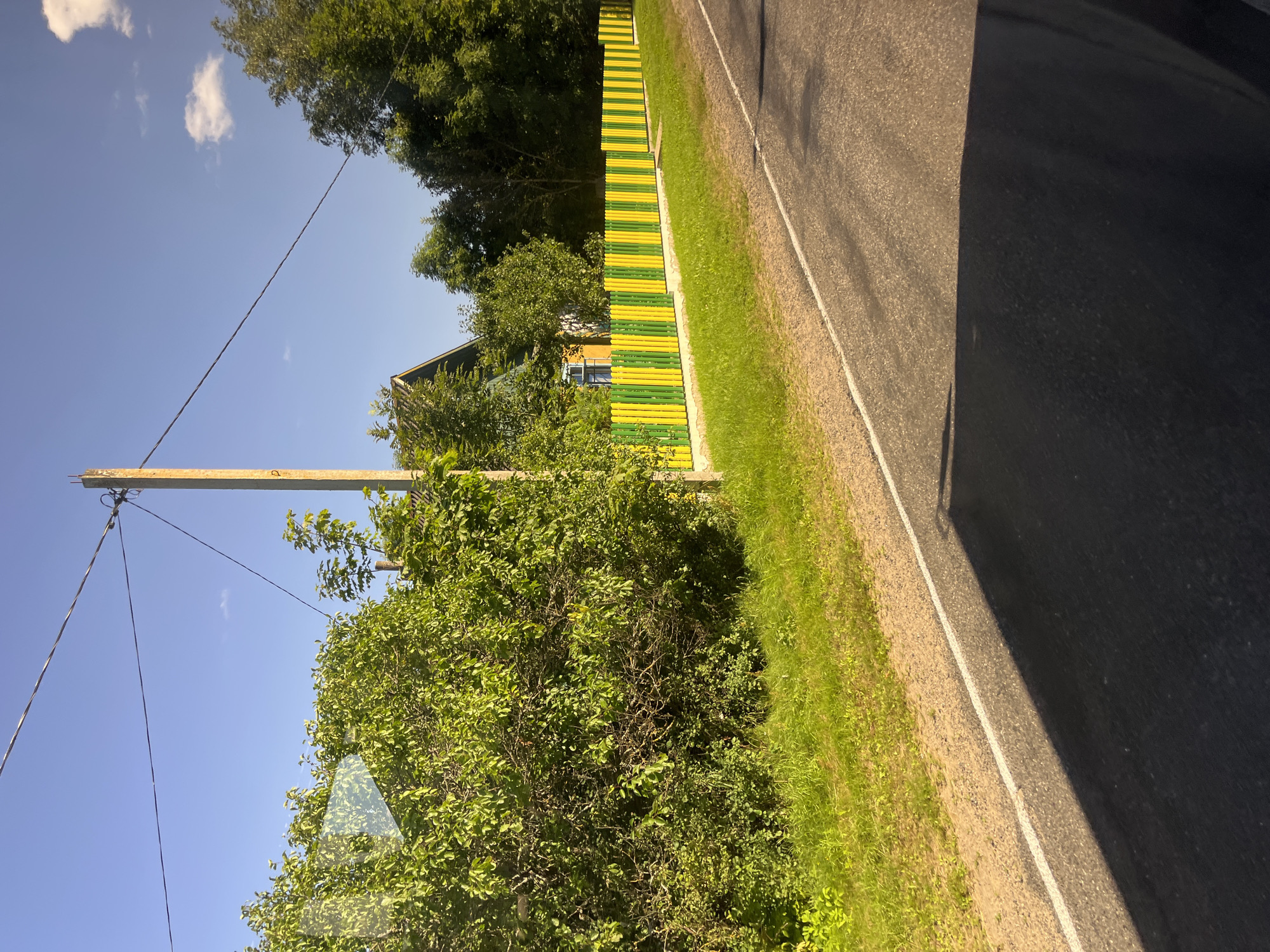 The image depicts a scene along a rural road, likely within the Мядзельскі сельскі Савет of Belarus, under a clear blue sky with scattered white clouds. On the left, a dense, vibrant green hedge or tree line dominates the foreground and middle ground, with a concrete utility pole positioned centrally. Beyond the foliage, the peak of a dark-roofed house is visible. A distinctive yellow and green striped wooden fence runs horizontally across the middle ground, bordering the property. The road itself occupies the lower portion of the image, with a white dashed line marking its edge and a prominent shadow cast across its surface, suggesting it is a sunny day. The environment appears to be a tranquil, sunlit rural setting. There are no people visible in the image.