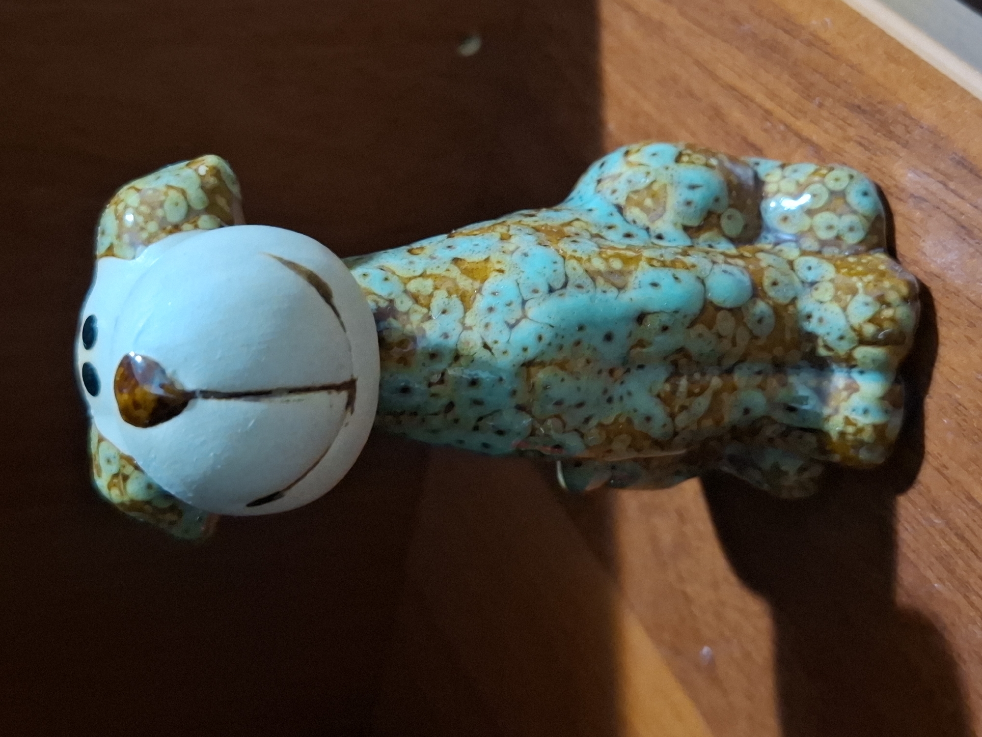 A ceramic dog figurine is the subject of this close-up, eye-level shot. The figurine has a light blue head with a wide, painted smile, black dot eyes, and a brown nose with a dark brown vertical line extending from it. The ears are light blue with brown speckles. The body is predominantly light blue with brown speckles and some areas of brighter yellow-brown. The figurine is sitting with its legs tucked under it. It is positioned on a wooden surface, likely a table or shelf. The background is dark and out of focus, suggesting it might be a wall or a darker shelf behind the figurine. The lighting is soft, casting a shadow to the left of the figurine. There is no indication of the location being in Almaty, Kazakhstan, or any other specific city.