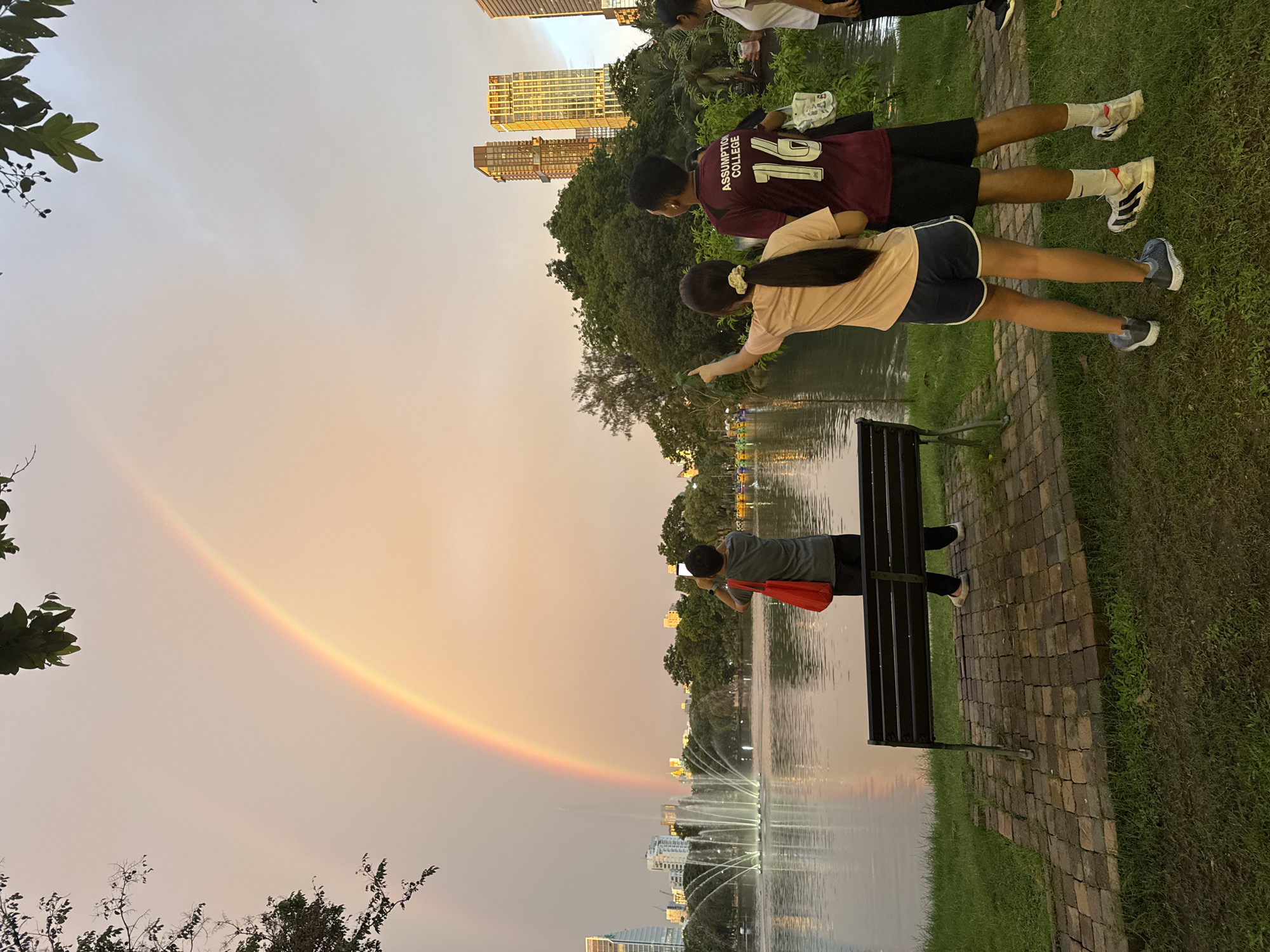 In a park setting in Bangkok, Thailand, a group of people are gathered near a lake with fountains. The sky displays a vibrant rainbow, suggesting it may have recently rained or is about to. The time of day appears to be late afternoon or early evening, with the sky showing warm, sunset hues.
In the foreground, a person in a grey t-shirt is standing on a paved path, holding a red bag and taking a picture with their phone towards the lake and the rainbow. Next to them, a bench is situated on the grass.
Further back, two individuals are standing on the grass. One person is wearing a maroon t-shirt with "ASSUMPTION COLLEGE" and the number "12" on the back, along with black shorts. They are looking towards the lake. Beside them, a person in a peach-colored t-shirt and blue shorts is reaching out towards the grassy area.
In the background, tall buildings are visible, indicating an urban environment. Trees and lush green grass make up the park's landscape. The water in the lake is calm, with fountains actively spraying water. The overall mood seems relaxed and appreciative of the natural phenomenon.