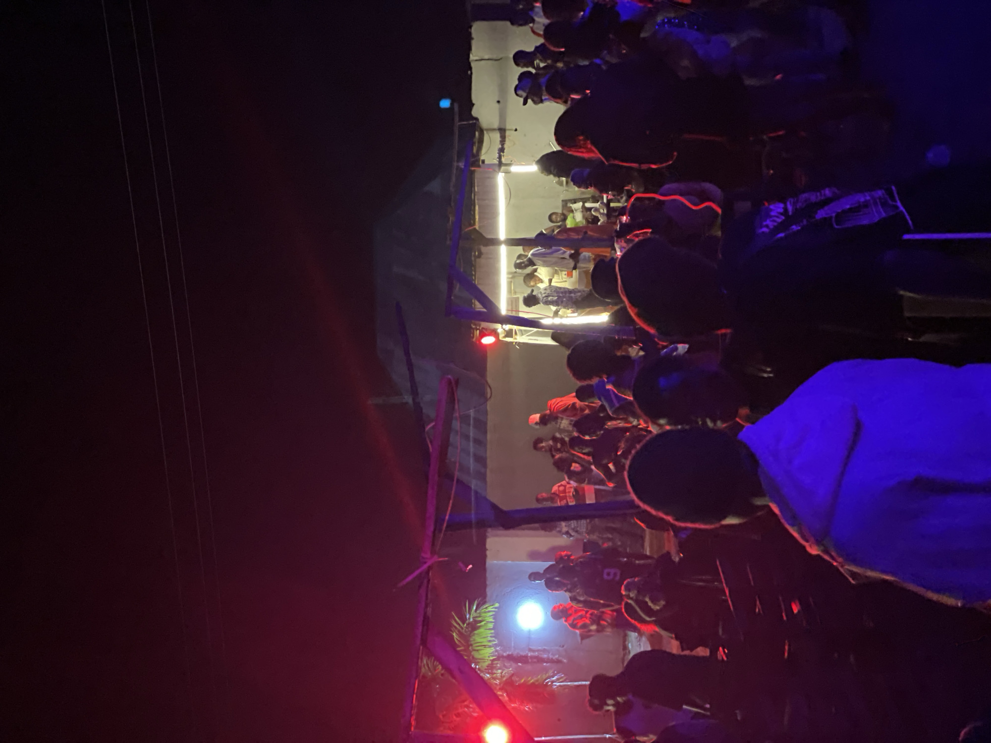 The image depicts an outdoor gathering at night in the city of Durumi, Nigeria, illuminated by red and blue stage lighting. A diverse crowd of approximately 30-40 individuals is present, engaged in various activities such as socializing and observing a performance or event.
In the foreground, a person wearing a blue hoodie is facing away from the camera, illuminated by the blue light. Behind them, other individuals are visible, some with their backs to the viewer. The midground features a raised platform with a roof structure, possibly a stage or stall, from which a bright white light emanates. Several people are gathered around this area, some appearing to be interacting with vendors or performers. Red lights are also prominent, casting a red glow on the scene.
The environment appears to be a somewhat informal outdoor space, with visible power lines overhead. Palm fronds can be seen to the left, suggesting a tropical or sub-tropical location. The overall atmosphere is lively and suggests a festive or celebratory event. The ground appears to be uneven, and there are some visible structures in the background, including what looks like a building with corrugated roofing. The lighting creates a dynamic and somewhat blurred effect, characteristic of night-time photography at an event with moving subjects.