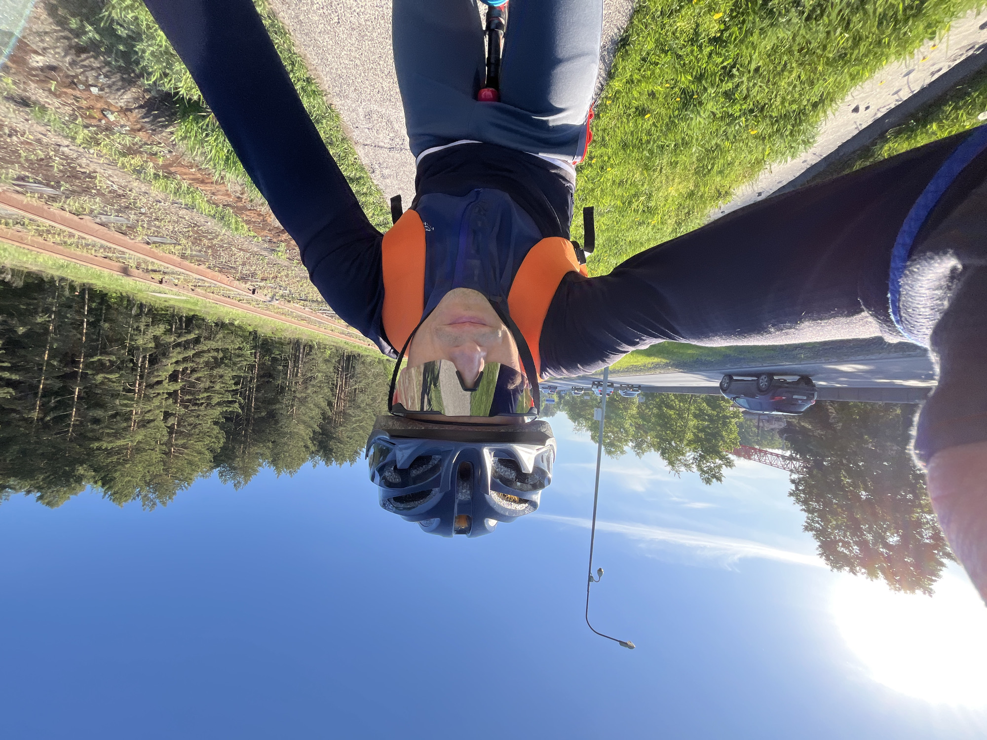 A selfie taken by a person on a bicycle. The person is wearing a cycling helmet and reflective sunglasses. They are wearing a dark blue, long-sleeved shirt with orange accents. The view in the background includes a lake reflecting trees, a road with cars, and greenery. A bridge is visible in the background. The location is Chaikovsky, Russia.