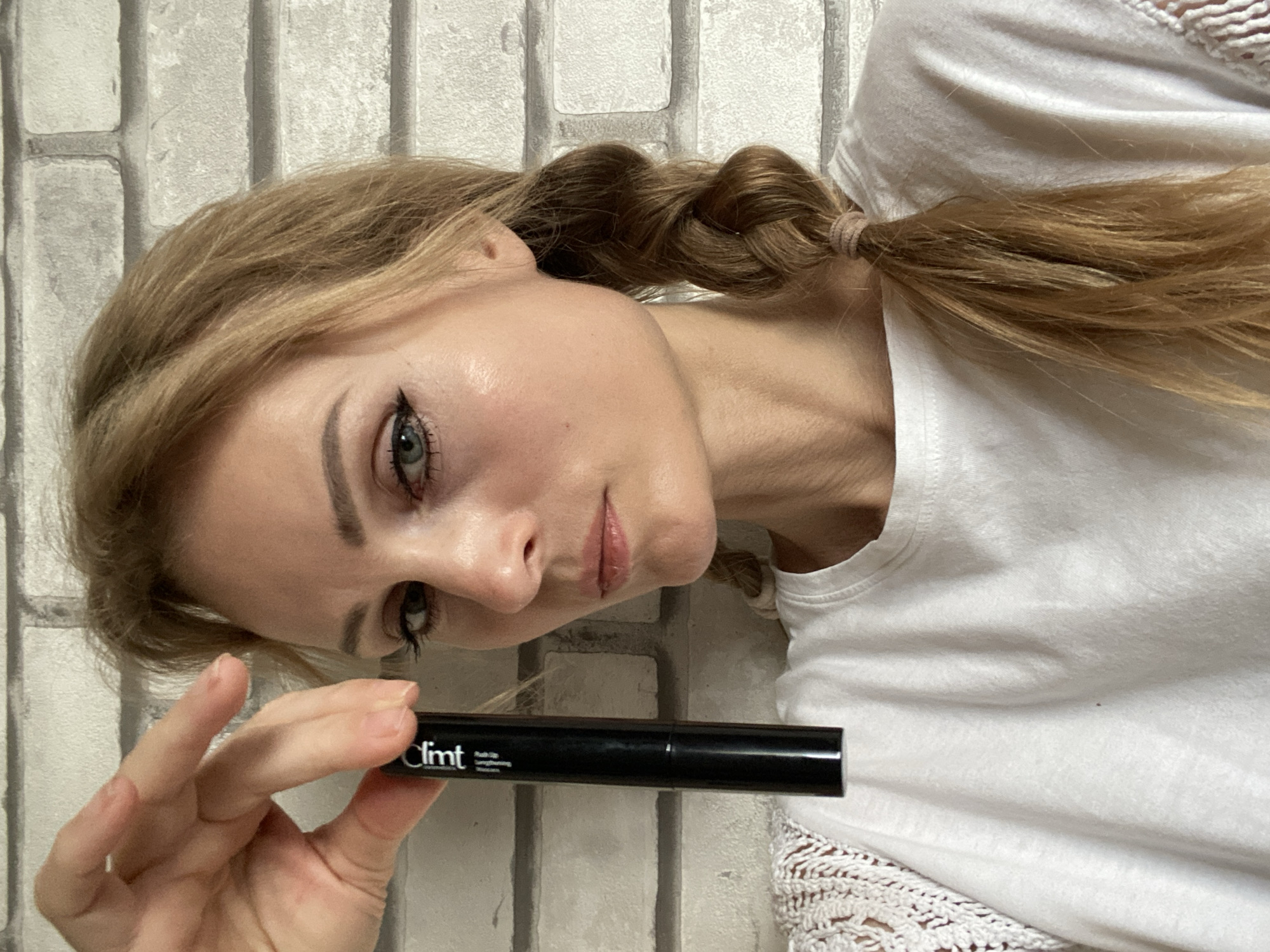 The image shows a medium shot of a young Caucasian woman with blonde hair in a braid, holding a black tube of mascara. She is wearing a white t-shirt with lace detailing on the sleeves. Her makeup includes eyeliner and mascara. The background is a white brick wall. The scene appears to be indoors, likely in a residential setting. The location is identified as Миханавіцкі сельскі Савет, Belarus.