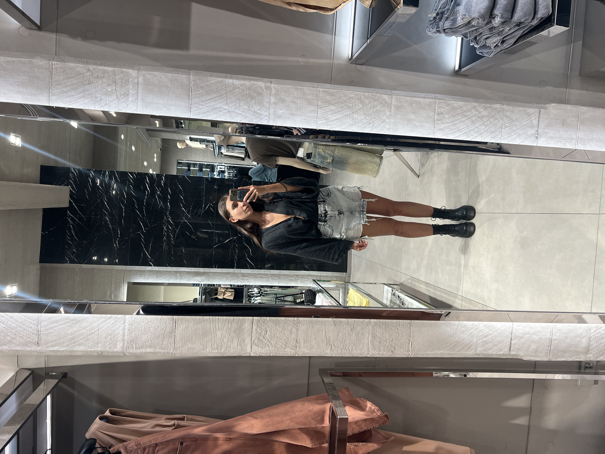 The image shows a woman taking a picture of herself in a mirror inside a retail store. The woman is wearing a dark top, a denim mini skirt, and black boots. She is holding a smartphone in her right hand.
The background of the image shows a typical retail store environment. There are clothes racks with clothing, shelves with folded garments, and marble wall panels. The store's flooring appears to be light-colored tile.
There are no specific details to indicate the location in the city of Unknown, Unknown.