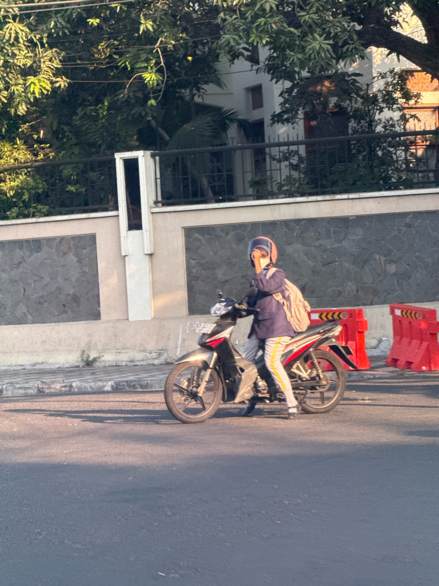 The image depicts a person riding a motorcycle on a paved road in Surabaya, Indonesia. The individual, wearing a blue jacket, light-colored pants with yellow stripes, a helmet, and a backpack, is positioned in the center of the frame. Their right hand is raised towards their face, possibly adjusting a mask or sunglasses. The motorcycle is silver and red. In the background, a light-colored wall with dark stone accents forms a boundary, and beyond that, lush green trees and a building are visible. Red traffic barriers are placed to the right of the motorcycle. The lighting suggests it is either early morning or late afternoon, with long shadows cast on the road. The scene is urban, characterized by the road, wall, and traffic barriers.