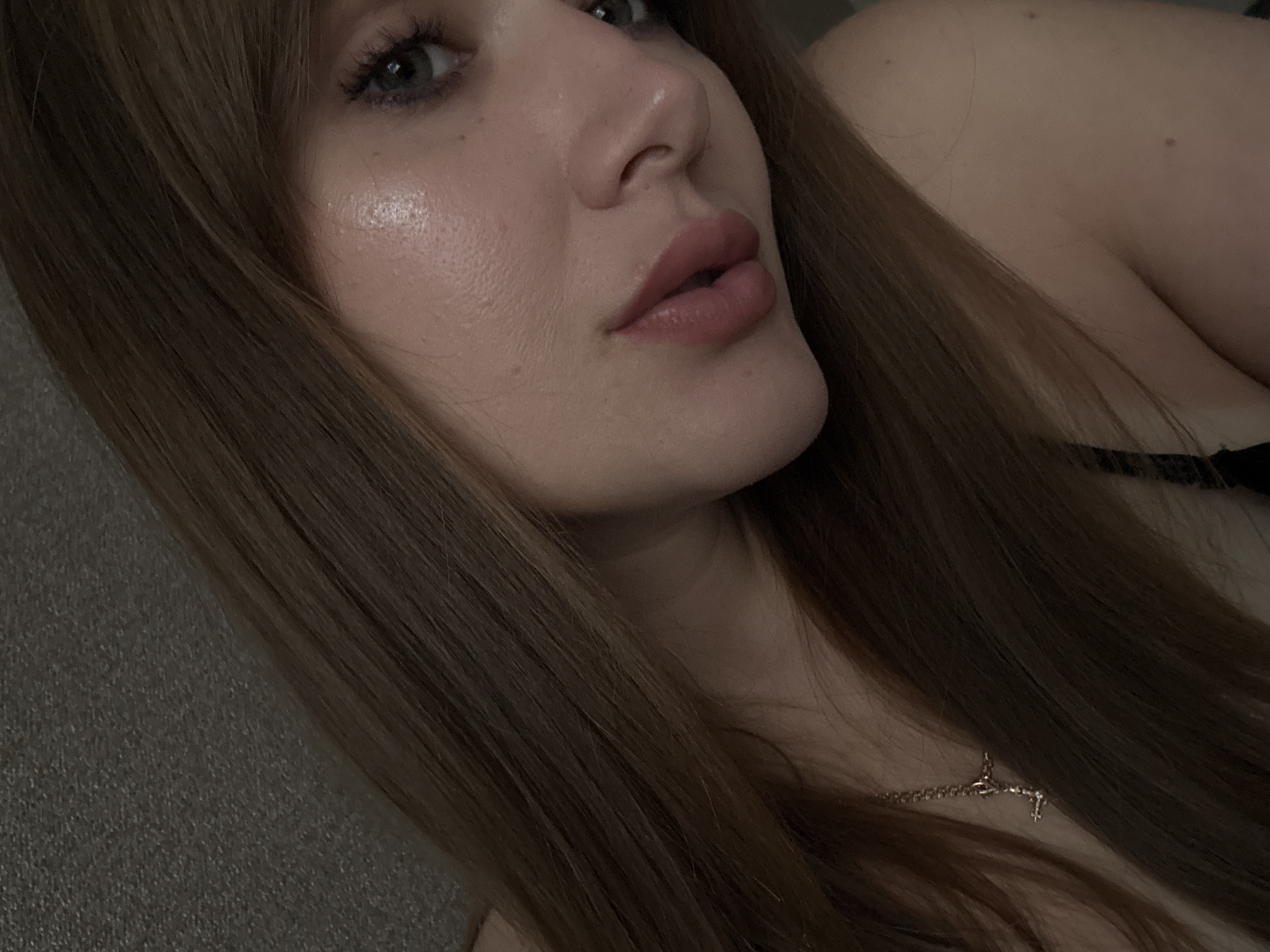 This close-up portrait captures the right side of a person's face and upper body in a relaxed, intimate setting. The subject, likely female, has long, straight, medium-brown hair that frames her face and falls over her shoulder. Her right eye, appearing grey or blue, is visible with subtle eye makeup. Her nose is prominent, and her full lips are tinted with a reddish-pink lipstick. The skin on her cheek shows a natural texture and a soft highlight.
The person is positioned as if lying down, with her gaze directed slightly upwards. A slender golden chain necklace with a small pendant, possibly a cross, rests on her upper chest, peeking through her hair. A dark-colored garment, appearing to be a bra strap or top, is visible near her shoulder.
The lighting is soft and dim, suggesting an indoor environment, possibly in the evening or at night. The background is indistinct and appears to be a dark, textured fabric, likely part of a couch or bedding, contributing to the casual and calm atmosphere. There are no overt location cues visible in the image itself to directly indicate Chaikovsky, Russia. No text is visible.