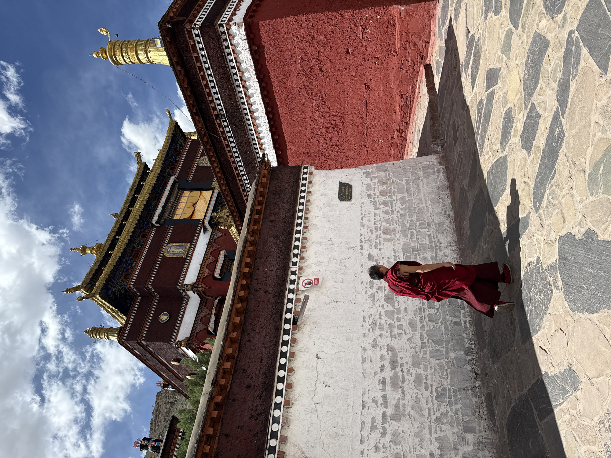 A wide, medium-shot, low-angle photograph shows a red-robed monk walking along a stone path adjacent to a building in the city of 城西街道, China. The monk faces away from the viewer. The exterior of the building is ornate, red and brown, with gold embellishments and a multi-tiered roof. The sky is blue with scattered clouds. The stone path is made of various sizes and colors. A portion of the building's wall is red, while another section is white with black and white patterned trim. A sign is affixed to the white wall. The image appears to be rotated.