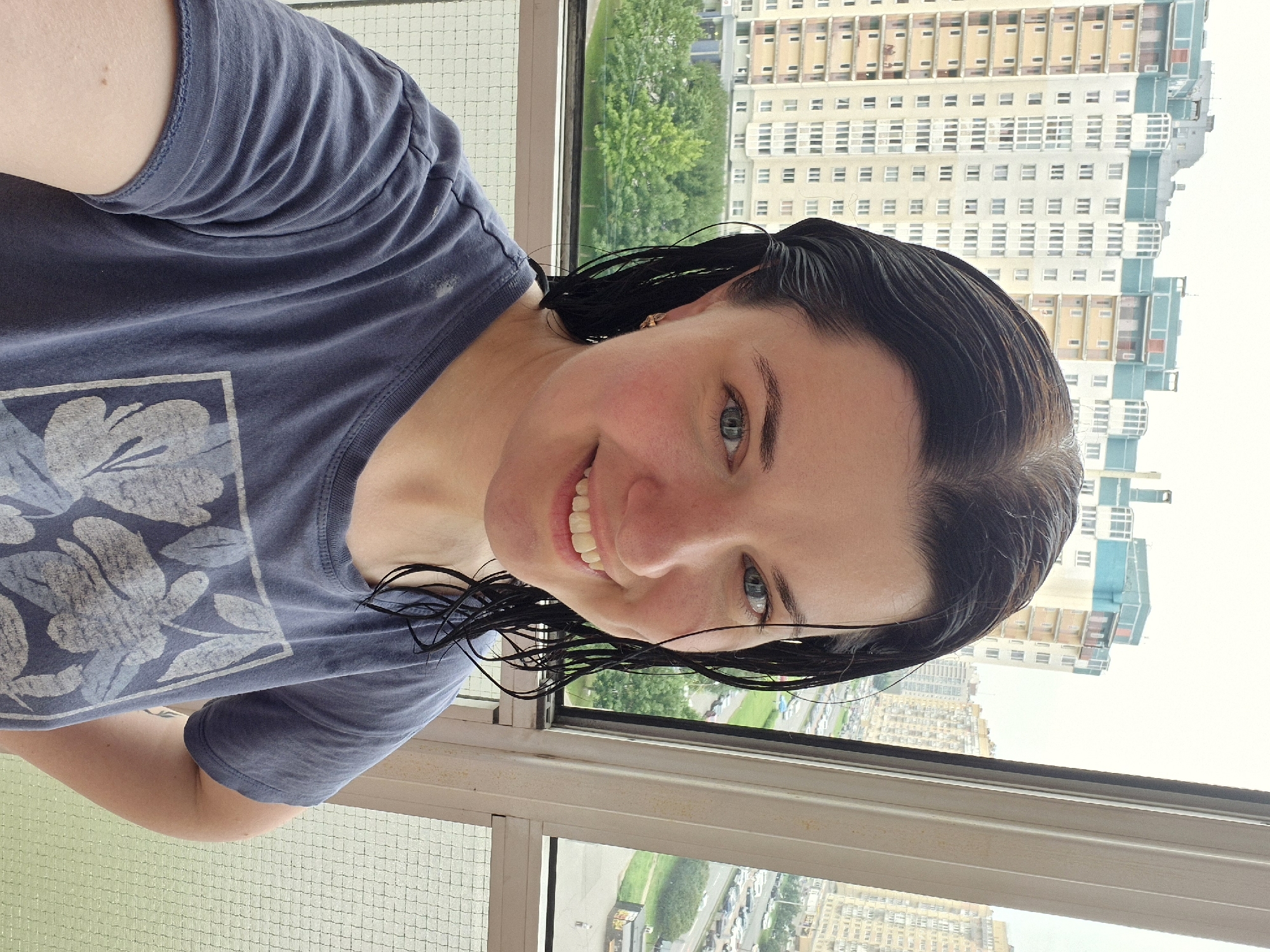 A woman with wet, dark hair and blue eyes smiles at the camera. She is wearing a navy blue t-shirt with a floral print. The woman is standing on a balcony with a city skyline visible in the background. The buildings appear to be apartment complexes. The weather is overcast, and the trees are lush and green. This scene is likely Saint Petersburg, Russia.