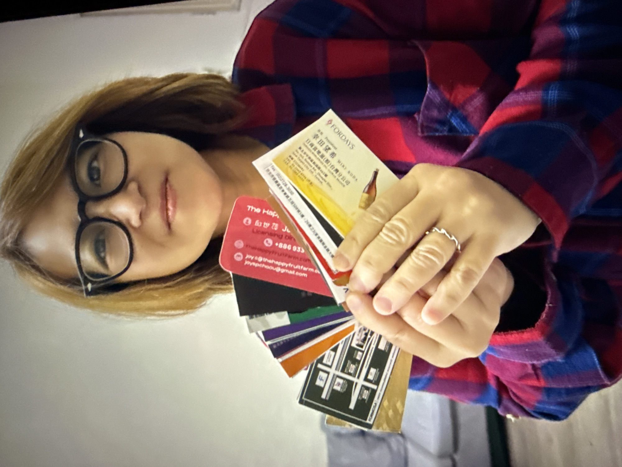 A woman wearing glasses and a red and blue plaid shirt holds a fan of various business cards and identification cards. The background is a plain, light-colored wall, and a portion of a couch is visible on the left side of the frame. The woman is looking directly at the camera with a neutral expression. The business cards are diverse in color and design, with some featuring text and logos that are partially obscured. The scene appears to be indoors, likely in a residential or office setting. There is no specific indication of the location being in Taipei, Taiwan.