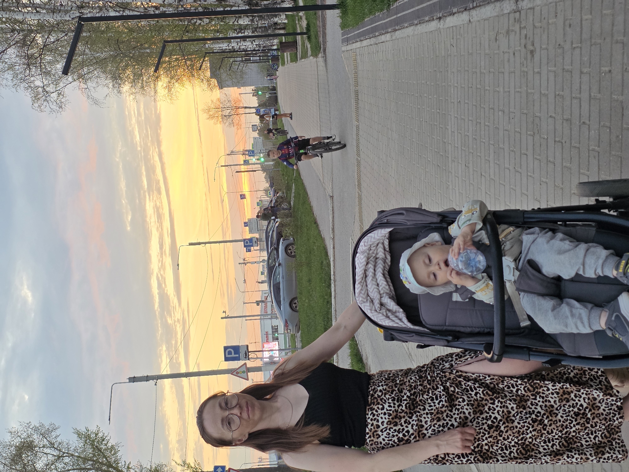The image captures a scene in the city of Chaikovsky, Russia, on what appears to be a sidewalk or pedestrian pathway. A woman with long brown hair, wearing glasses, a black top, and a leopard-print skirt, stands next to a baby stroller. Inside the stroller, a baby is lying down, holding a water bottle.
In the background, the sky is a mix of light blue and golden colors, suggesting either a sunrise or sunset. Further away, there is a road with traffic signs and parked cars. A young person on a bicycle is riding on the sidewalk in the same direction as the woman and stroller. Utility poles and trees line the edge of the road. The streetlights are visible, which indicates that it is near dusk or dawn. The pavement of the sidewalk is made of interlocking brick.