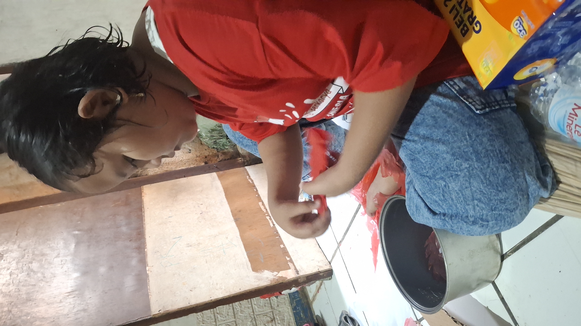 A young child, likely female, wearing a red t-shirt and blue jeans, is sitting on the floor. The child is looking down and appears to be engaged in an activity involving red plastic material. In front of the child, to the left, is a metallic pot containing some dark-colored items. To the right of the child, a box with yellow and blue packaging is visible, along with a plastic water bottle. The environment appears to be indoors, with a tiled floor and a wooden structure in the background.