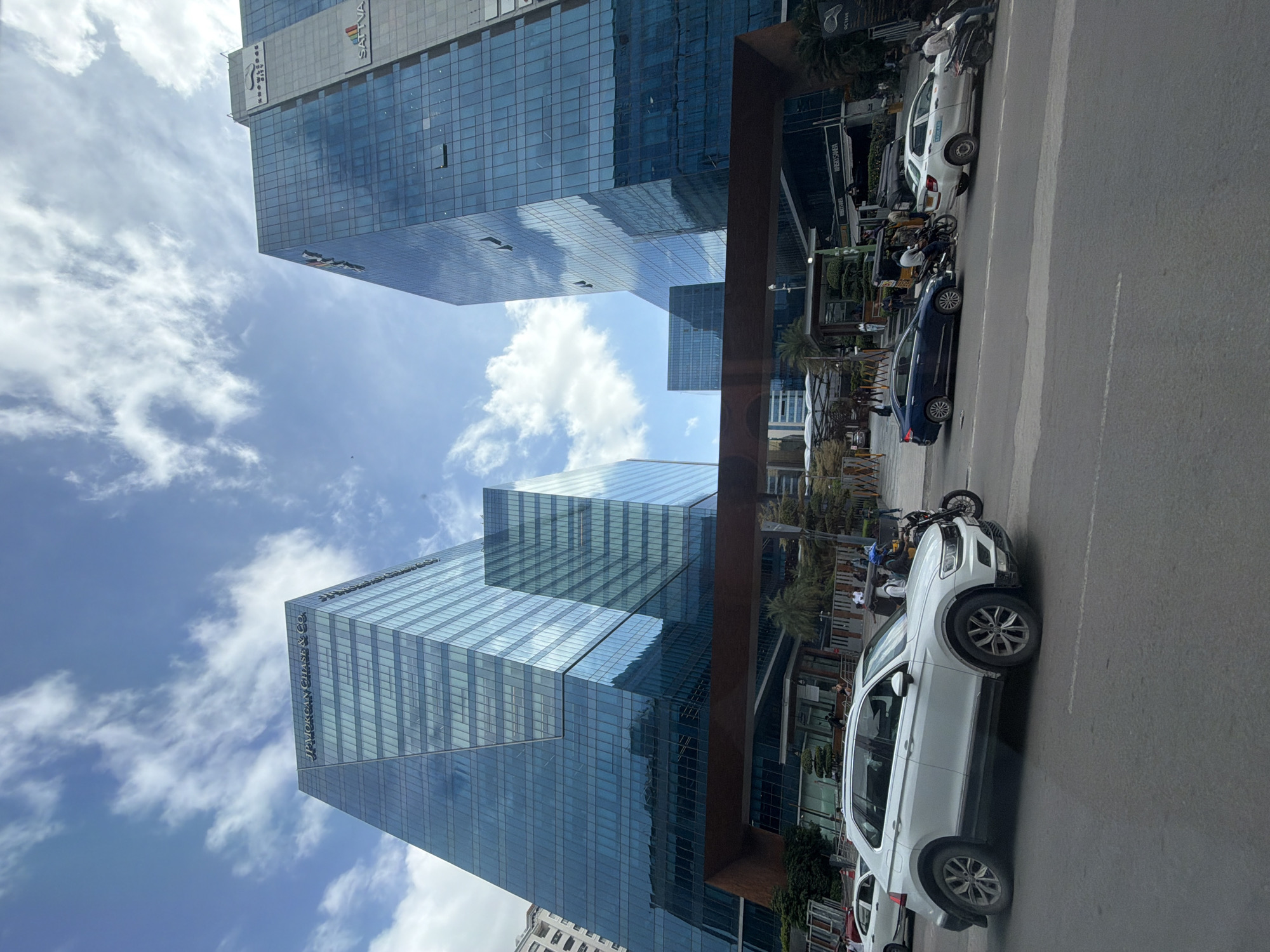 The image captures a street view in Hyderabad, India, featuring the JPMorgan Chase & Co. building and the Sattva Knowledge City. Dominating the scene are two modern, glass-facade office buildings. The JPMorgan Chase & Co. building, on the left, is a striking geometric structure with a distinct angled design. The Sattva Knowledge City building, on the right, also boasts a sleek glass exterior and displays signage including the Sattva logo.
Below these imposing structures, a wide asphalt road is visible, bustling with traffic. A white SUV, likely a Volkswagen Tiguan, is prominently positioned in the foreground, moving along the road. Several other vehicles, including cars, a motorcycle, and an auto-rickshaw, are also present, indicating moderate urban traffic. Pedestrians can be seen walking on the sidewalks and near the entrance of the buildings. The environment appears to be a commercial or business district, with trees and landscaping adding a touch of greenery. The sky above is a mix of blue and scattered white clouds, suggesting daytime.