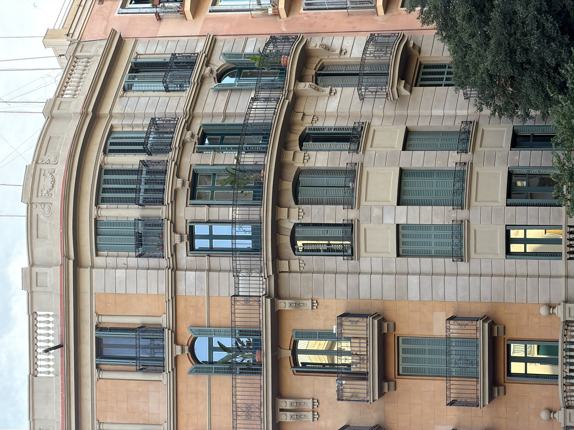 This image shows a close-up, straight-down view of the facade of a multi-story building in Barcelona, Spain. The building features a mix of architectural styles, with some sections exhibiting classical influences, such as columns and decorative carvings, while others have a more modern appearance with clean lines. The facade is predominantly beige and salmon-colored, with various windows and balconies.
The windows are mostly rectangular, some with green shutters, and others with reflective glass showing the sky. Several balconies with ornate black wrought-iron railings are visible, some adorned with potted plants, including a large palm-like plant. The building appears to be an apartment building or hotel, with multiple residential or commercial units.
The time of day is difficult to ascertain precisely due to the overcast sky, but the lighting suggests it could be daytime. The weather appears to be cloudy. The image captures the intricate details of the building's construction, from the textured stonework to the delicate metalwork of the balconies. There is no visible text in the image. The overall impression is one of architectural richness and urban density characteristic of Barcelona.