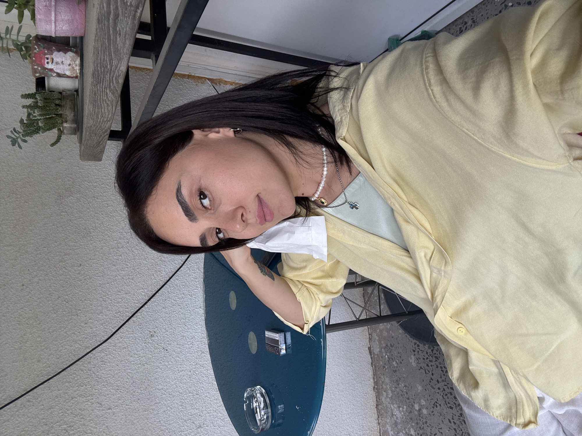 A woman with dark hair and brown eyes is sitting at a teal table outdoors. She is wearing a light yellow button-up shirt over a mint green shirt, and a pearl necklace with a pendant. She is holding a white tissue to her face. On the table are an ashtray with cigarette butts and a pack of cigarettes. Behind her, a wall with a black cable runs diagonally. To the right of the woman, a black metal shelf holds potted plants, one of which is decorated with a snowman. The floor is a grey concrete patio. The scene appears to be on a balcony or patio in Tbilisi, Georgia.