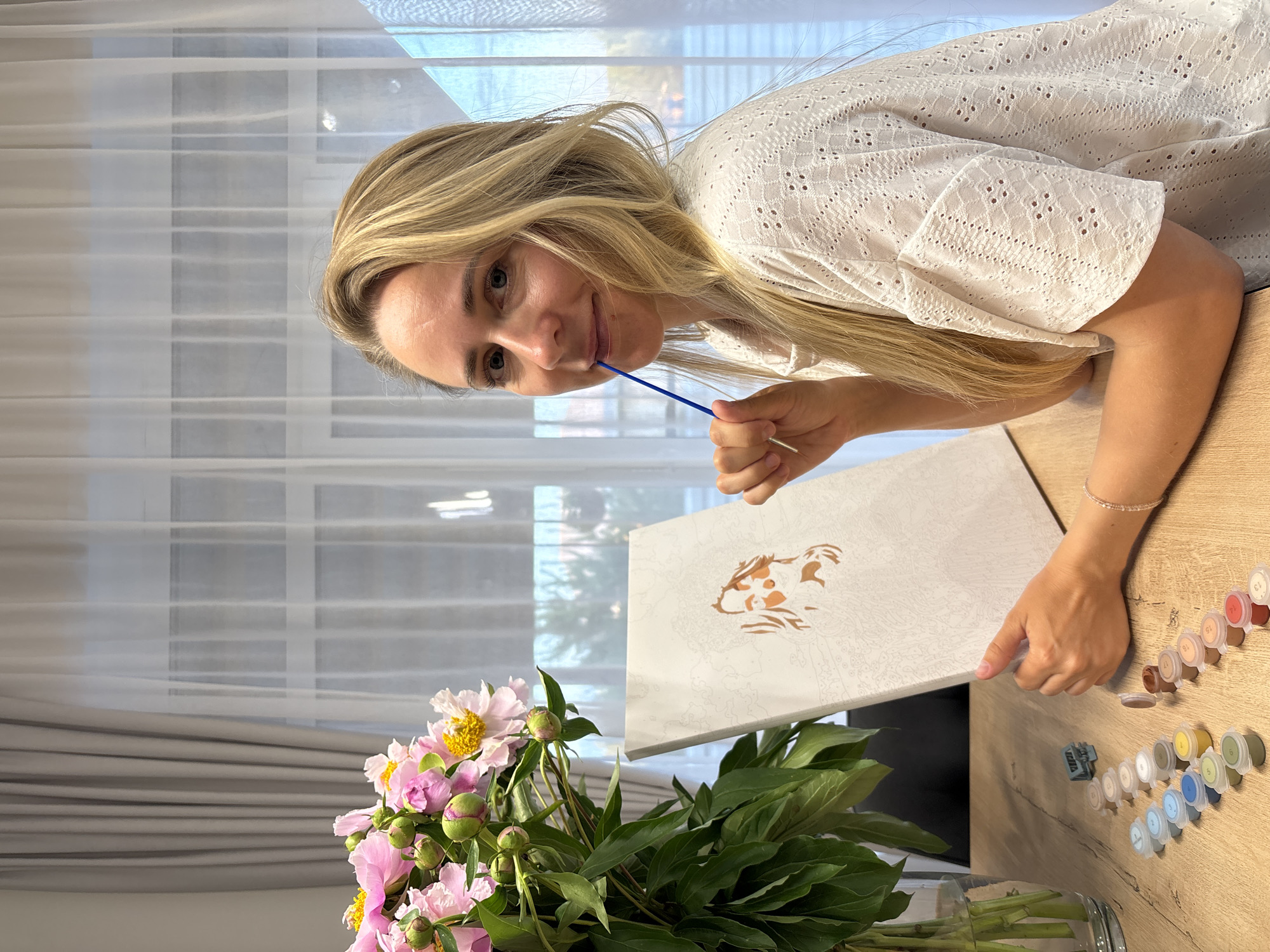 A blonde woman is sitting at a wooden table, with a bouquet of pink peonies in a glass vase to her left. In front of her is a paint-by-numbers canvas with an incomplete portrait of a woman. She is holding a blue paintbrush in her mouth and looking towards the camera. Various small paint pots are arranged on the table. The background shows sheer white curtains over a window. The scene appears to be indoors, possibly a home. The location in Chaikovsky, Russia, cannot be determined from the image.