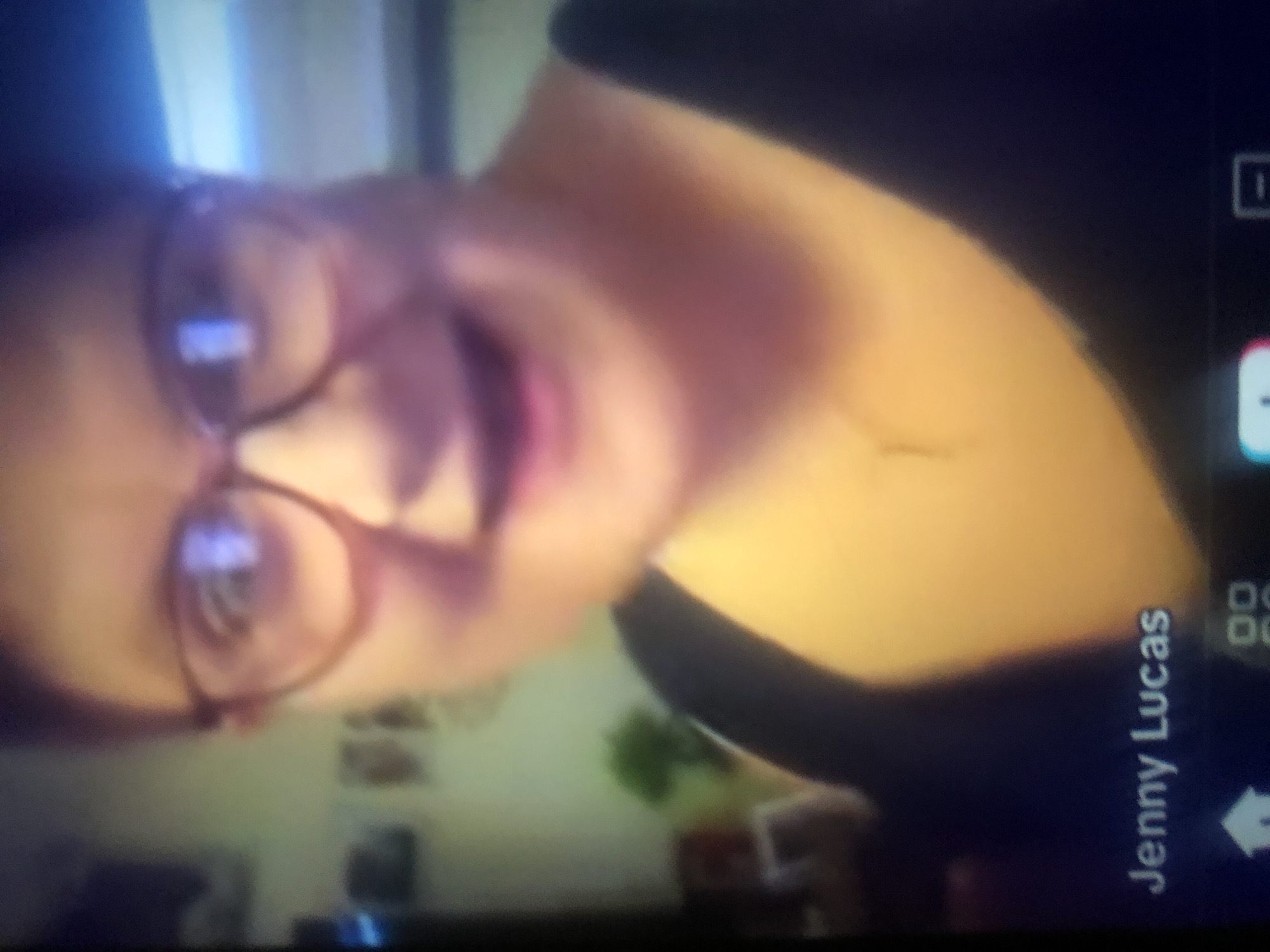 A blurry, close-up video still shows a woman, identified as Jenny Lucas, with glasses and a dark top. She is smiling and looking at the camera. The background is out of focus but appears to be an indoor setting with some shelves or decorations visible. The bottom of the image displays interface elements, suggesting it is from a video call or a screen recording. The location in the city of Howayi, Nigeria is not discernable from the image.