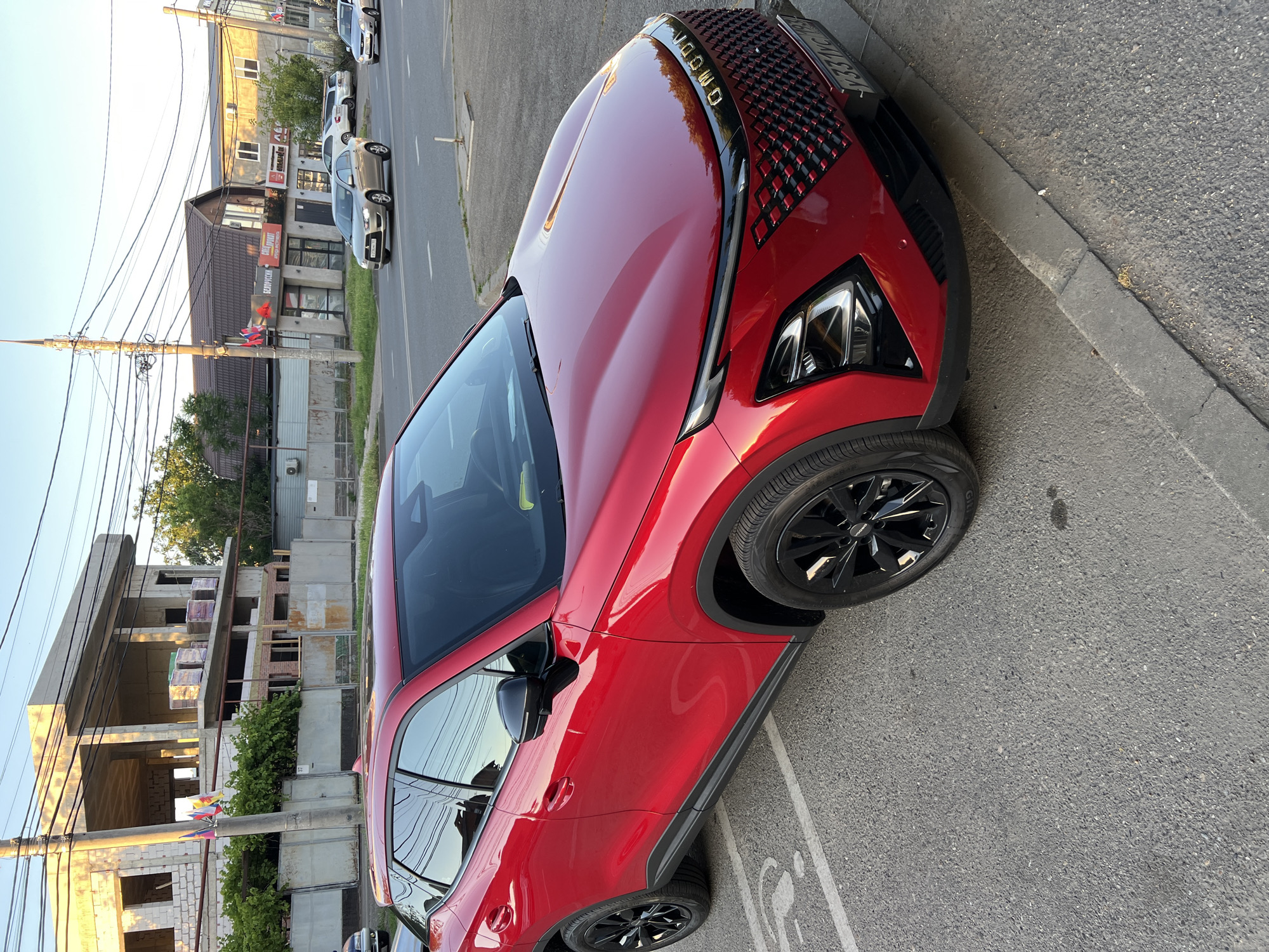 The media file shows a red car parked on an asphalt surface next to a sidewalk. The car is a modern SUV with a black grille and black wheels. The sun is casting long shadows, suggesting it is either early morning or late afternoon. The scene appears to be urban, with buildings and power lines visible in the background. One building under construction is prominent on the left, with concrete frames and stacked materials. Further down the street, other cars are parked and moving, indicating traffic. There is some vegetation, including trees and bushes, growing along the street and near the buildings. The overall impression is a typical street scene in a residential or commercial area. The license plate of the red car is partially visible, showing Cyrillic characters and numbers, consistent with a Russian license plate. The location context is Krasnodar, Russia. No people are clearly visible in the image.