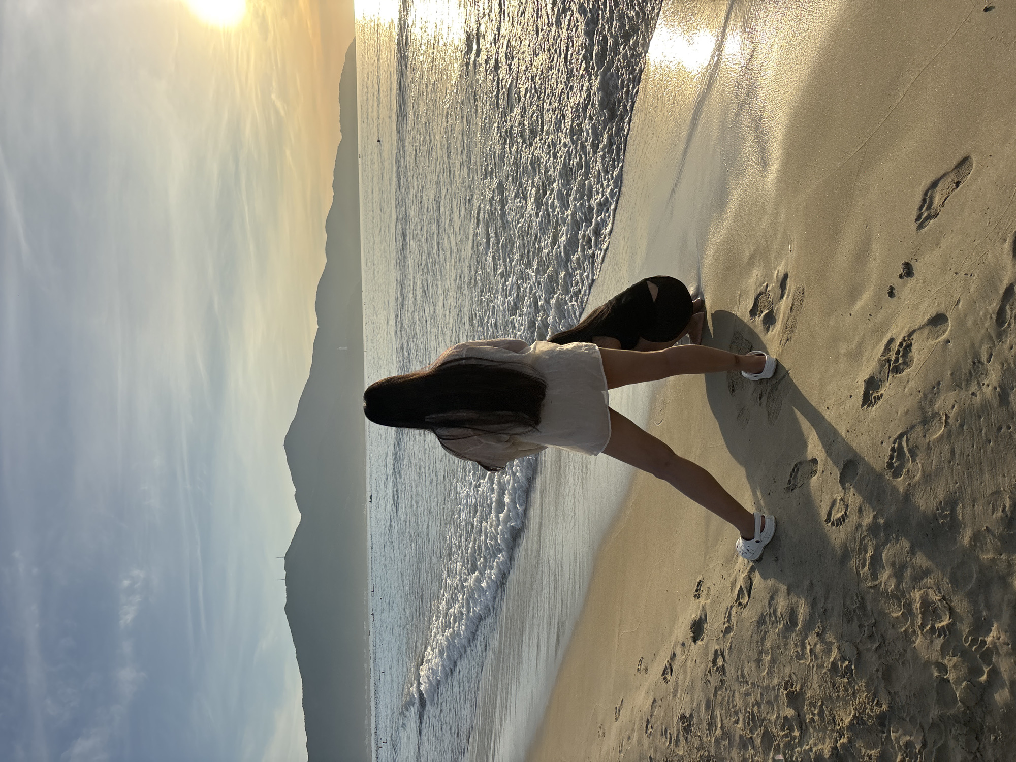 A woman with long dark hair stands on a sandy beach in Da Nang, Vietnam, facing the ocean. The sun is setting, casting a warm golden glow on the water and the sky. Mountains form a silhouette in the distance. The woman is wearing a light-colored shirt and shorts and white slip-on shoes. Her shadow stretches across the sand, marked with footprints. Another person, wearing dark clothing, is crouched down near her. Waves gently roll onto the shore.