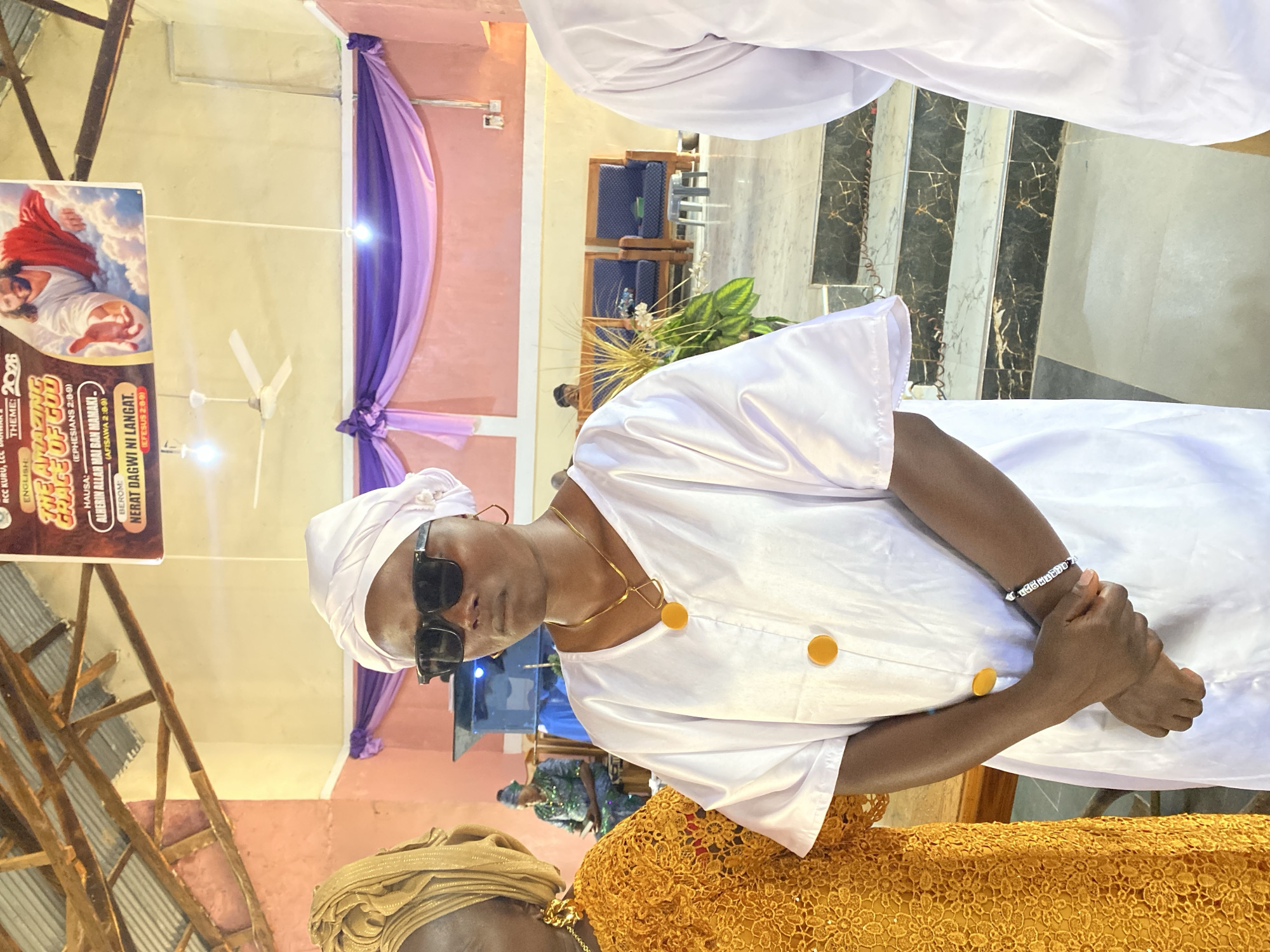 The image depicts a woman in a white outfit and head wrap, wearing sunglasses and a gold necklace. She has her hands clasped. Part of another woman in a gold outfit is visible in the lower part of the frame. The background includes purple drapes, ceiling fans, and what appears to be a church interior. A banner reads "RCC Kuru, The Amazing Grace of God", indicating a location in Kuru Dungus, Nigeria. The banner contains additional text in Hausa and Berom.