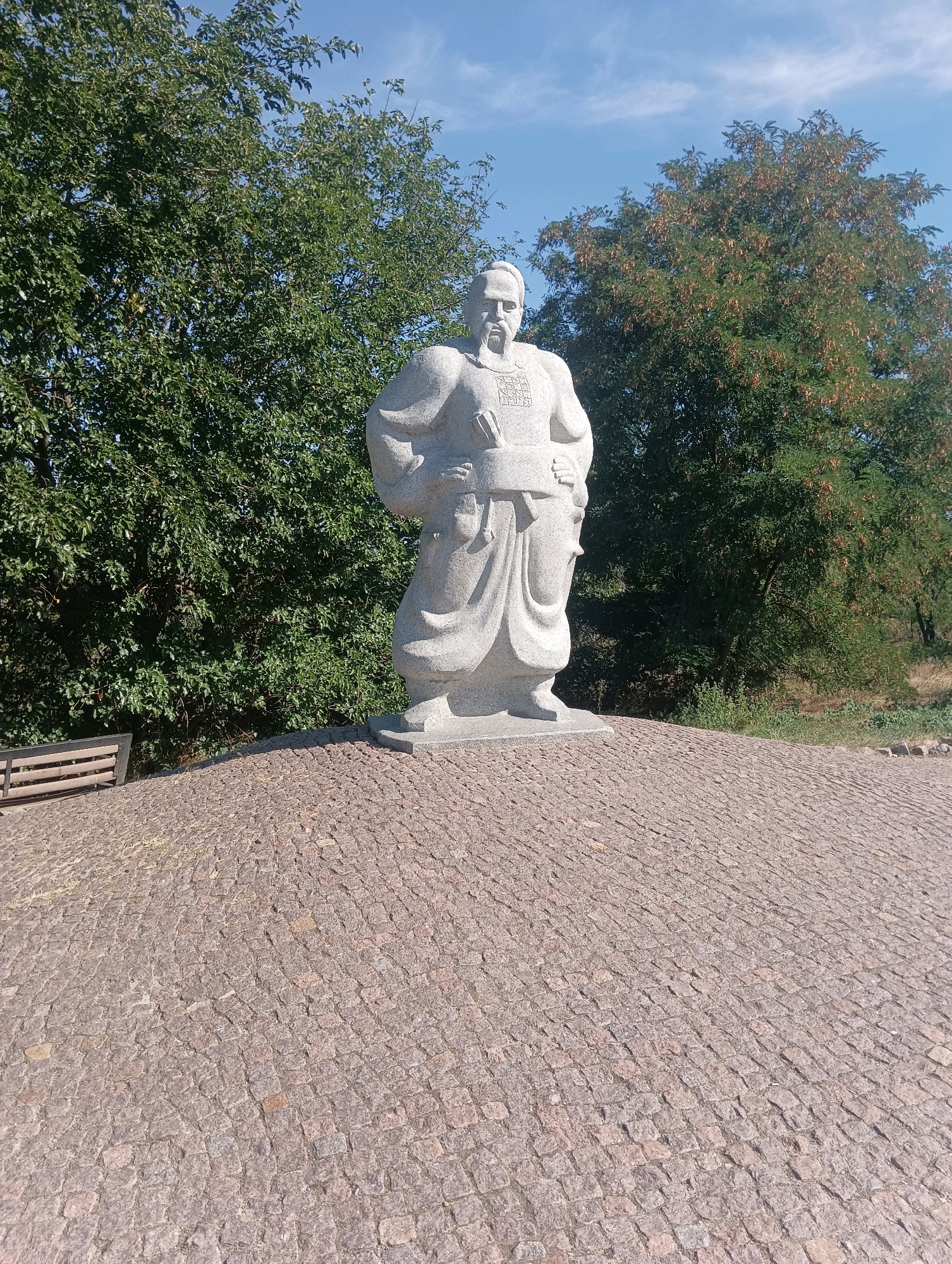 An outdoor, eye-level, full shot captures a stone statue of a man with a beard and a traditional outfit. The statue is positioned centrally on a cobblestone pedestal that slopes downwards towards the viewer. Behind the statue, a lush green tree fills the left side of the frame, while another tree with dry seed pods occupies the right side. The background consists of a clear blue sky with scattered white clouds. A portion of a wooden bench is visible to the left of the statue, near the base of the pedestal. The overall lighting suggests a sunny day.