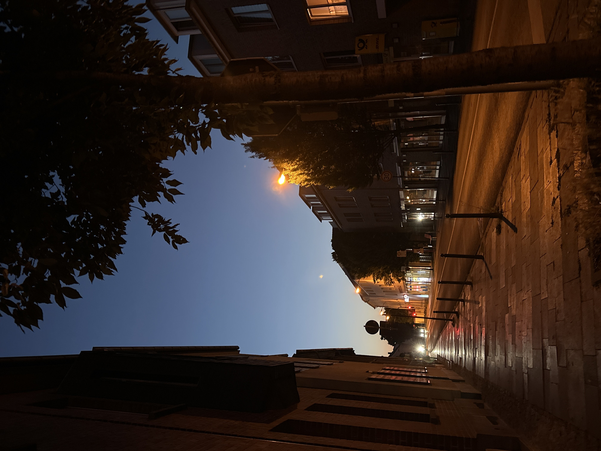 The image depicts a street scene in Krefeld, Germany, during twilight. On the left, a dark brick building with multi-paned windows lines the sidewalk. The sidewalk is paved with light-colored stones, wet and reflective, with black bollards marking its edge. A street with a single white lane marker runs alongside the sidewalk. Several streetlights illuminate the scene with a warm orange glow. On the right, buildings with illuminated windows and storefronts are visible. A tree with dark foliage stands prominently in the foreground on the right, obscuring part of the buildings behind it. A yellow DHL package dispenser is attached to one of the buildings. Traffic lights and road signs are present. The sky is a deep blue, transitioning to a lighter hue near the horizon. There are no people visible in the image.