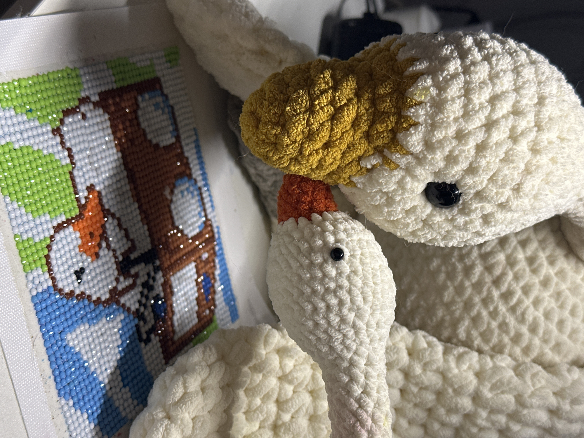 The image shows two crocheted plush geese next to a diamond painting of a child wearing blue boots and standing outside of a brown house with green trees. The larger goose has a white body, yellow head, and black eyes. The smaller goose is white with a pink neck and an orange beak. It is unclear if the geese are interacting. The environment is domestic and suggests a craft project in progress. It is impossible to determine the location without additional information.