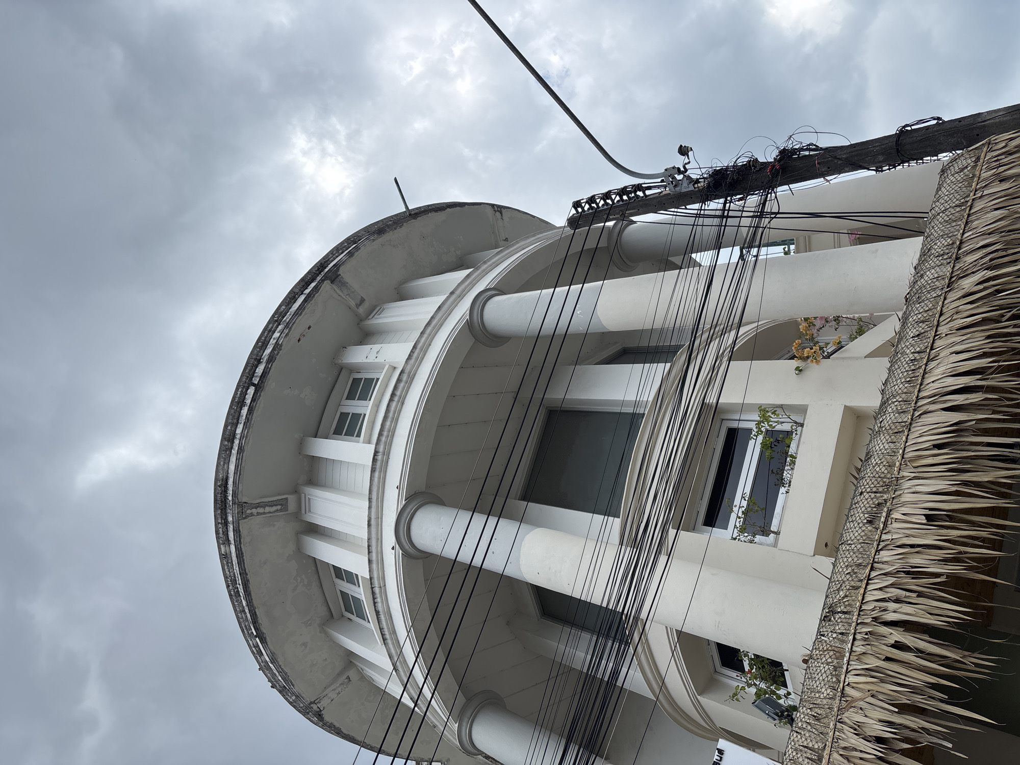 A low-angle shot of a cylindrical building in Phuket City Municipality, Thailand, under a cloudy sky. The building features white columns and windows on multiple tiers. A utility pole laden with tangled black wires is visible near the building's edge, partially obscured by a section of thatched roofing. The building's facade appears aged, with visible wear and discoloration.