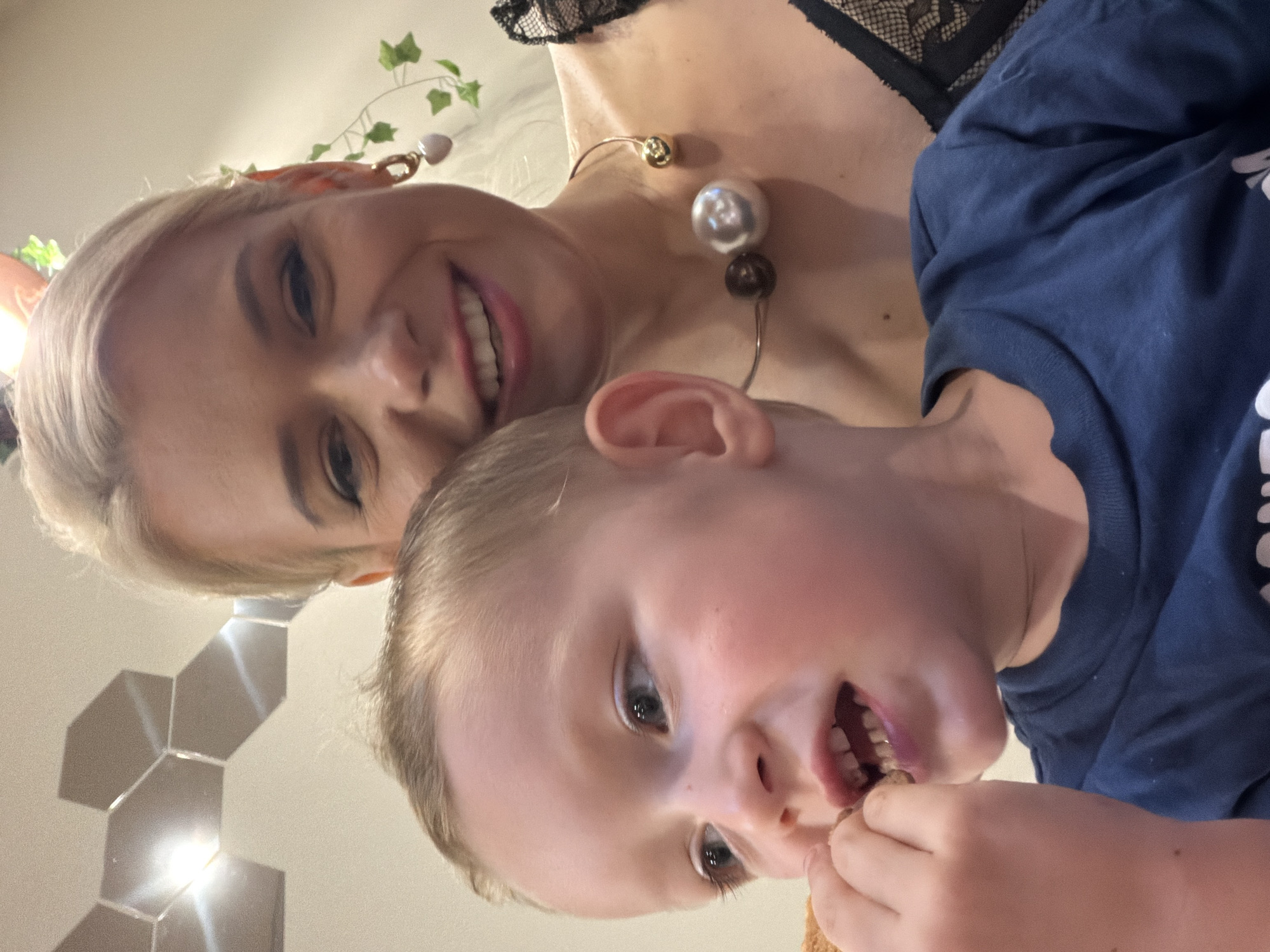 The image is a selfie taken indoors, likely in a home setting in Sochi, Russia, as indicated by the location context. The perspective is close-up and angled, suggesting the photo was taken by one of the subjects.
The primary subjects are a woman and a young child. The woman has blonde hair and is smiling brightly. She is wearing a necklace with several large, spherical beads in varying colors (gold, silver, and dark brown) and a black lace garment. A small, decorative green vine with leaves and a pink charm is visible in her hair.
The child, who is positioned closer to the camera and appears to be in the foreground, has blonde hair and is looking directly at the camera. The child is wearing a dark blue long-sleeved shirt. The child's mouth is open, and they are holding a small, brown object, possibly a piece of food, in their hands and bringing it to their mouth.
The background is soft and out of focus, but it appears to be an indoor environment with light-colored walls and some reflective hexagonal decorative tiles on the left side of the frame, which catch the light. There are no clear indications of the time of day or weather conditions. The overall mood appears to be casual and intimate, possibly a moment of shared connection or play between the mother and child. There is no visible text in the image.