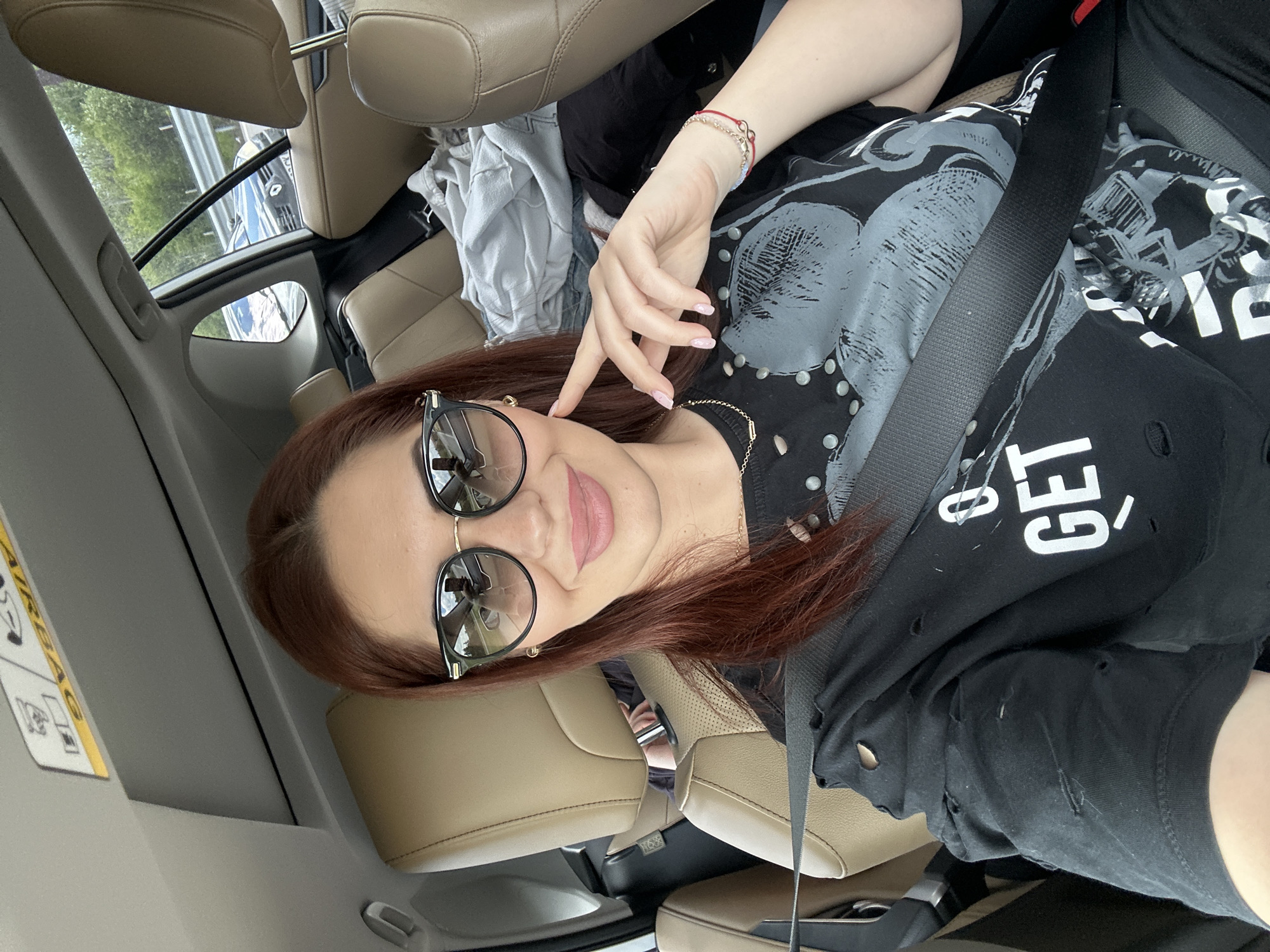 A woman with auburn hair and black round-frame sunglasses is sitting in the passenger seat of a car, wearing a black t-shirt with white lettering. She is holding her right hand up near her face, and a seatbelt is fastened across her torso. The car's interior is beige leather, and there is a pile of clothing behind her in the back seat. The location is likely Lozen, Bulgaria, based on the available information. The environment suggests a car interior, and the subject is a woman in the passenger seat.