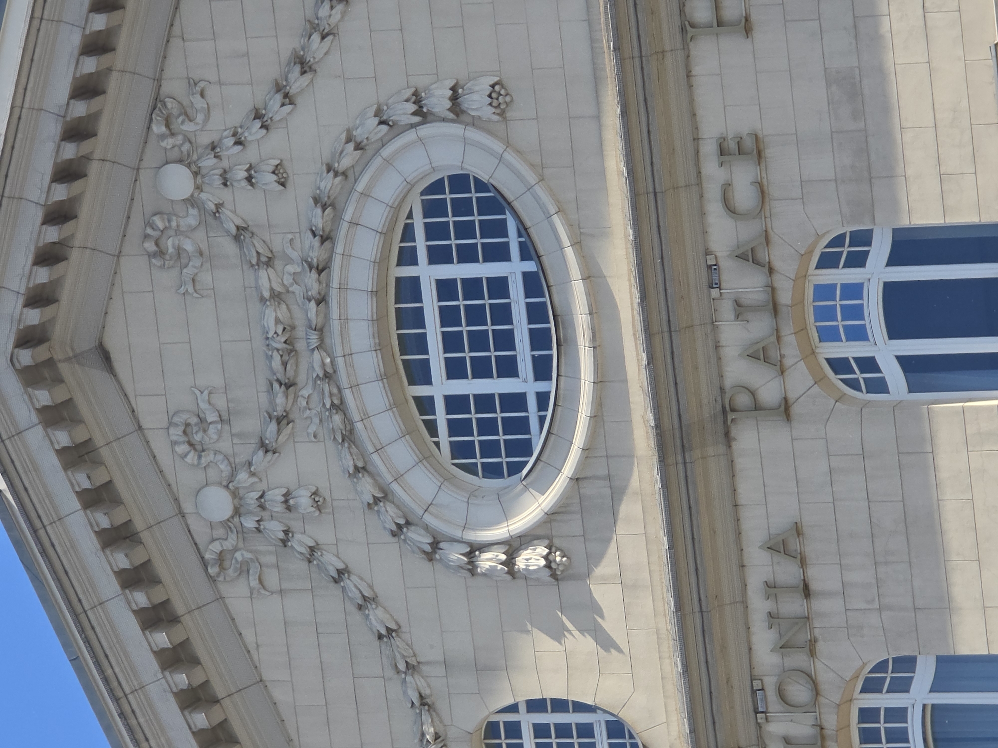 The image shows a section of a building facade, featuring architectural details such as an oval window with a grid pattern and decorative floral elements carved into the stone. The building's name, "IONIA PALACE", is vertically inscribed on the right side of the facade. Based on the name "Ionia Palace" and the architectural style, the building is likely located in Warsaw, Poland. The facade is light-colored, possibly made of stone or concrete. The sky is visible in the window, indicating it is daytime.