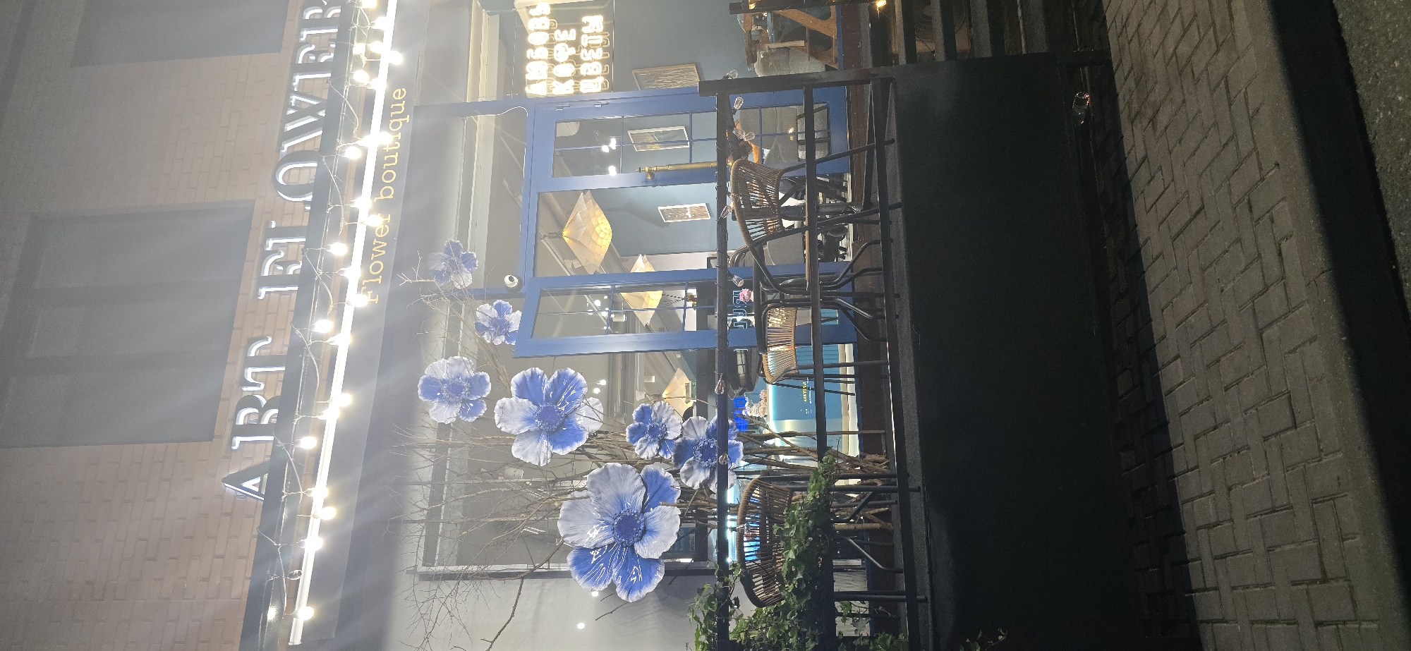 Here's a description of the image:
**Scene:**
The image shows the exterior of a flower boutique. The facade features blue trim around a large display window adorned with decorative blue flowers and branches. String lights are hung along the building's sign, which reads "ART FLOWERS boutique". Inside the shop, geometric light fixtures hang from the ceiling, and wicker chairs are visible. The building is located on a brick-paved street with a brick facade. The scene appears to be photographed at night.
**Location:**
The city of Grozny, Russia, cannot be determined from the image.