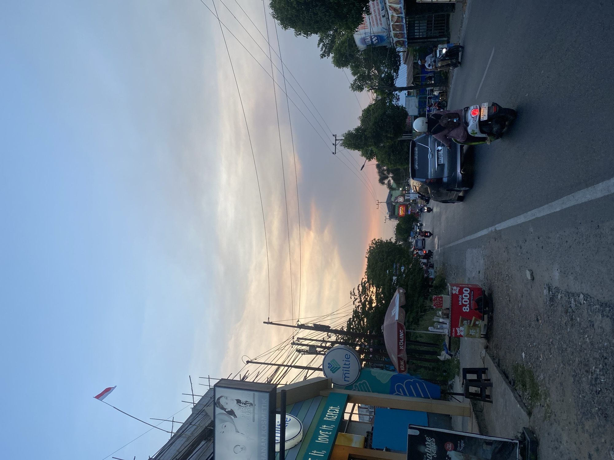 The image depicts a street scene in Banjarbaru, Indonesia, during the late afternoon or early evening, indicated by the soft lighting and colorful sky. A red and white Indonesian flag flies from a building on the left. The street is busy with traffic, including several motorcycles and a silver van. On the left side of the road, there are shopfronts with signage, including one for "Miltie" and another advertising cosmetic products. A vendor cart selling drinks is also visible. The right side of the street features more shopfronts, some with awnings, and trees lining the roadside. The sky is filled with a mix of light blue and soft, colorful clouds, suggesting a sunset. The road is paved and appears to be a main thoroughfare.