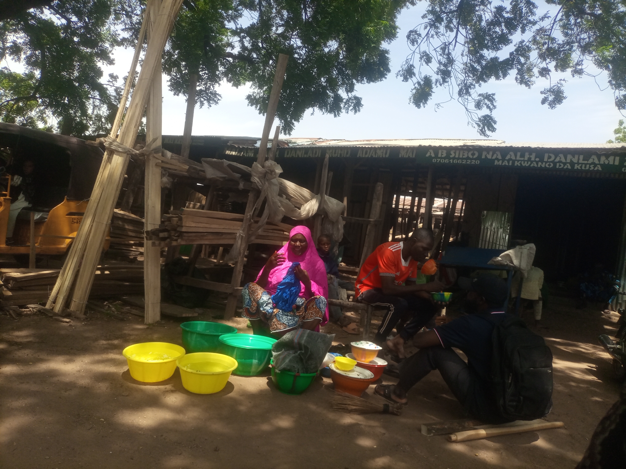 In Arawa, Nigeria, a scene unfolds under the dappled sunlight filtering through trees. Several individuals are gathered near a makeshift structure, possibly a market stall. A woman in a vibrant pink hijab and patterned clothing sits on the ground, holding a blue bag. Beside her, a child in a dark shirt looks on. To the right, a man in an orange t-shirt and dark pants is seated, holding a green bowl. Another man, wearing a dark t-shirt and a backpack, is also seated on the ground, facing away from the camera.
In the foreground, a collection of large plastic bowls, predominantly yellow and green, are arranged on the dusty ground. Some of these bowls contain water. Further back, stacked wood and bamboo poles suggest construction or a source of materials. The background reveals a corrugated metal roof structure with signage, some of which is legible, including "MAI... DANLADI MUHID... MAI... MAI KWANO DA KUSA" and a phone number "07061662220". The overall impression is of an outdoor marketplace or a working area in a rural or semi-urban setting.
