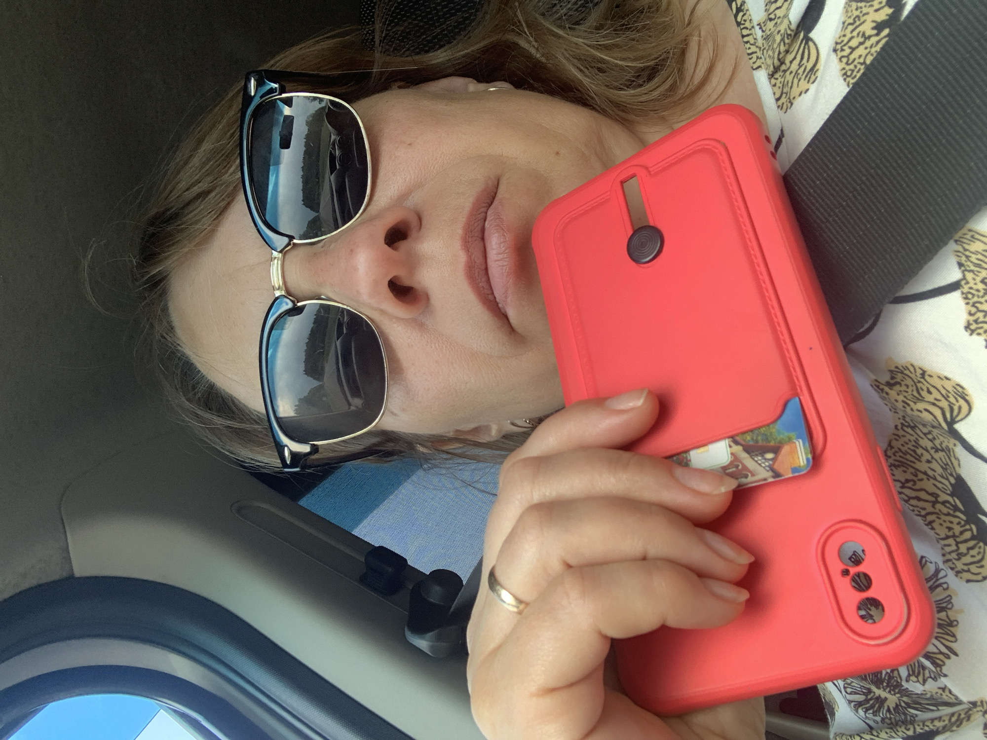 A woman in sunglasses is taking a selfie in a car. She is holding a red phone case with a card slot. The reflection in her sunglasses shows the road and trees. The car's interior is visible, including the seatbelt. The background is out of focus. The image appears to be taken during the daytime. There is no discernible information in the image to determine the specific location within Грозаўскі сельскі Савет, Belarus.