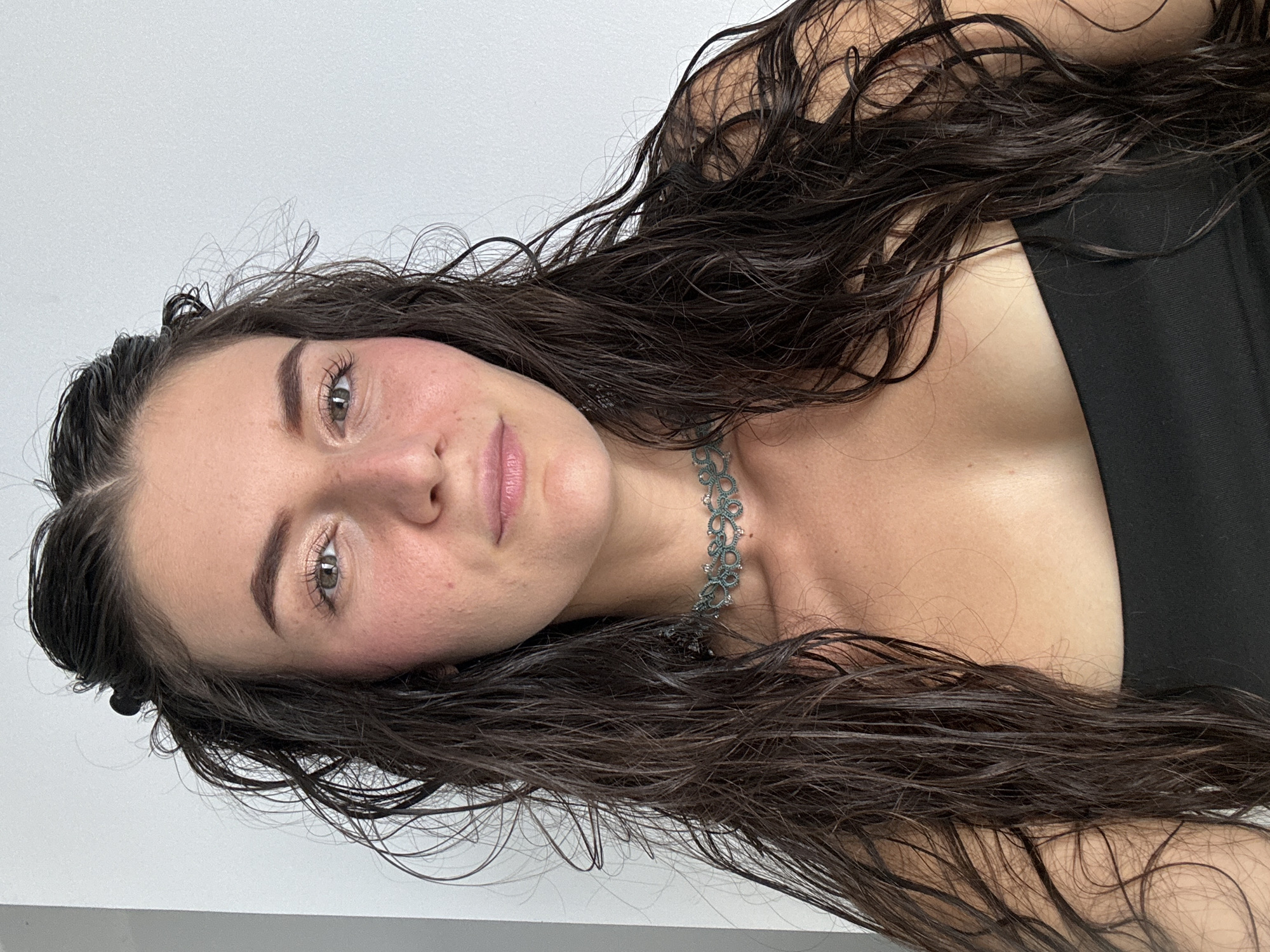 A light-skinned woman with dark brown, wet hair lies facing the camera. She has green eyes, light pink lips, and rosy cheeks. She is wearing a dark-colored top and a delicate green and silver choker necklace. The background is a plain, light grey wall. The location is undetermined as the image contains no contextual clues to suggest it was taken in Mũi Né, Vietnam.