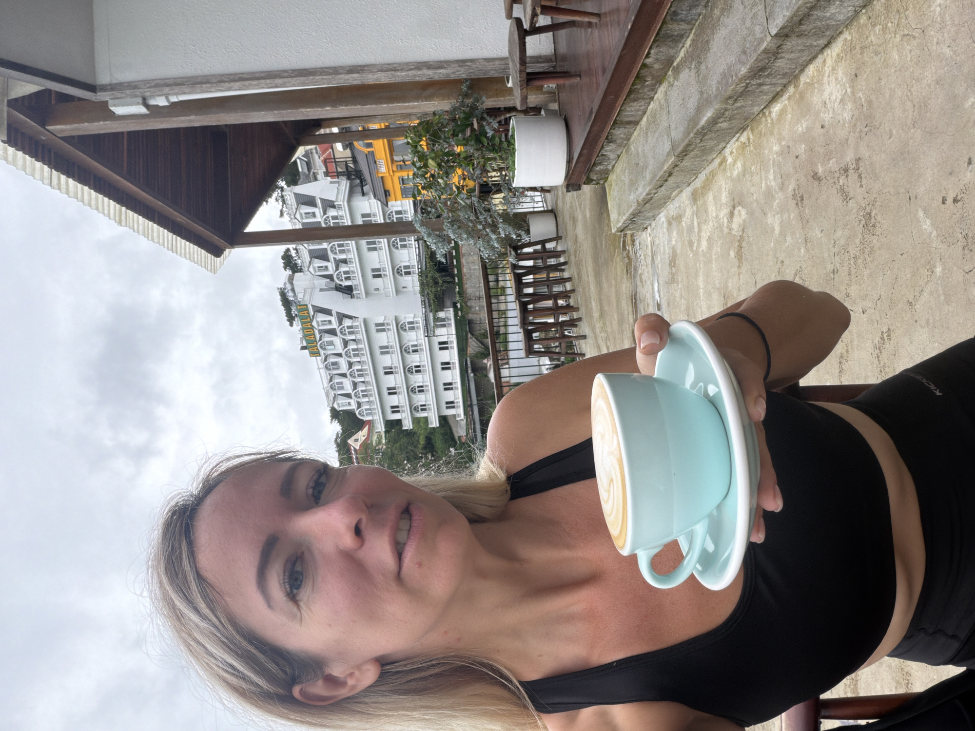 A woman with blonde hair is taking a selfie while holding a light blue cup of coffee with latte art, with a white building in the background. She is wearing a black sports bra and black leggings. The building in the background is the Dalat Palace Hotel, a historic landmark in Da Lat, Vietnam. The hotel is known for its French colonial architecture and has been a popular tourist destination since the early 20th century. The woman is standing on a balcony overlooking a town.