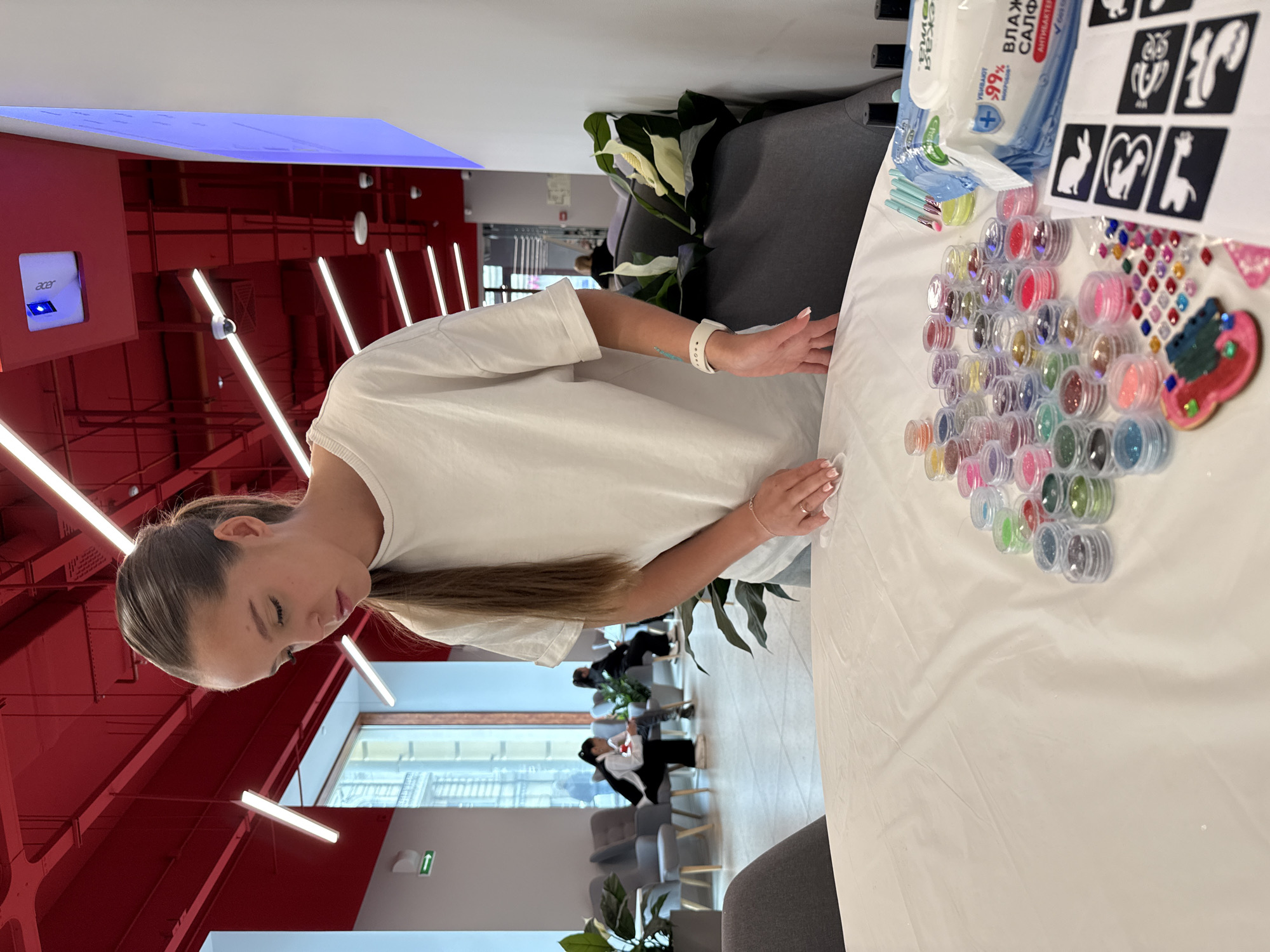 A woman with long brown hair, wearing a white t-shirt, stands in the foreground, looking down at a table. Her hands are positioned over the table, suggesting engagement in an activity. On the table, which is covered with a white cloth, a large collection of small, clear pots filled with various colors of glitter is arranged in an arc. Adjacent to the glitter are packages of wet wipes, a set of small brushes, and several stencils featuring animal shapes (such as a bunny, cat, owl, and giraffe) and other designs like a heart and a butterfly. Also visible are small, colorful gems or stickers. The woman wears a white smart watch on her left wrist and a ring on her left ring finger.
The setting is an indoor, modern space, likely an event area or lounge, in Vladivostok, Russia. The background features a distinctive red ceiling structure with linear white lights, and large windows revealing an outdoor view. Several individuals are seated in a lounge area with grey chairs and potted plants. A device labeled "Acer" is mounted on a red wall. The wet wipes package displays Cyrillic text including "ВЛАЖНЫЕ САЛФЕТКИ" (Wet Wipes), "АНТИБАКТЕРИАЛЬНЫЕ" (Antibacterial), "+99%", and "ВЫТИРАЮТ РУКИ" (Wipe Hands). The scene appears to depict a creative workshop or craft event, possibly for temporary glitter tattoos or similar art, occurring during the daytime.