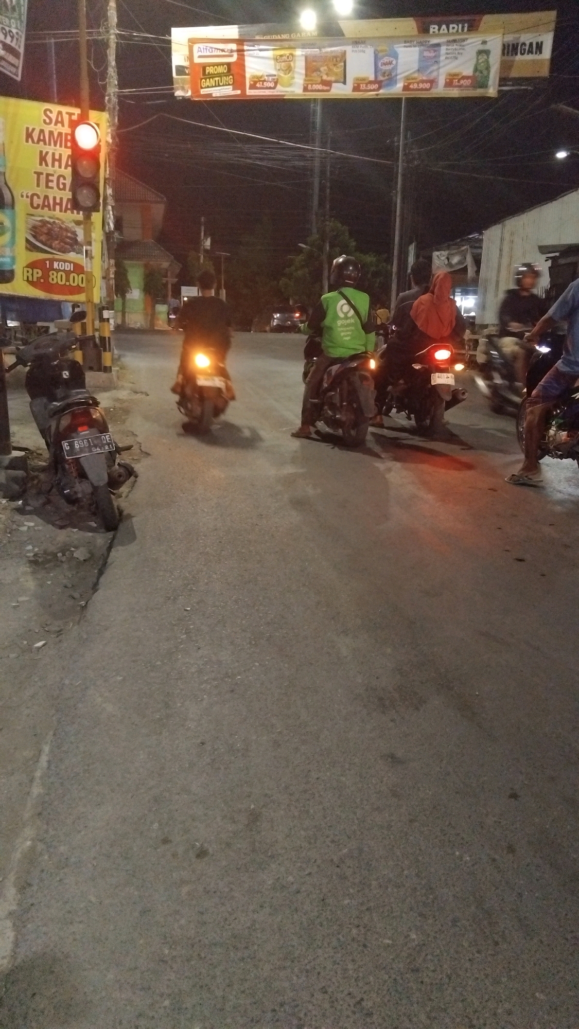 The image depicts a street scene at night in Tegal, Indonesia, likely near a commercial area, given the advertisements visible. A traffic light is illuminated red, indicating a stop. Several people on motorcycles are present. One person in a green Gojek uniform is on a motorcycle, facing away from the camera. Another person is riding on the back of this motorcycle. To the left, a parked motorcycle with license plate C 6961 OE is visible. Further down the street, other motorcycles and their riders are present, some with their headlights on. Overhead, banners display advertisements, including one for "Alfamart" and another with prices in Indonesian Rupiah. The road surface is asphalt, and there are overhead power lines. The overall atmosphere is that of a typical urban street at night with ongoing traffic.