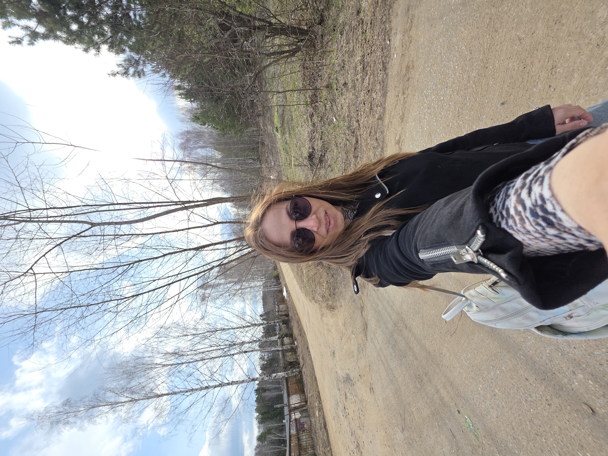 A woman with long, light brown hair is taking a selfie on a dirt road. She is wearing sunglasses, a black jacket, and blue jeans. She is holding a white backpack with a silver zipper. The location appears to be an unpaved road in a forest or park area, identified by the surrounding trees and vegetation. In the background, there is evidence of sparse residential buildings or fences. The photo was taken in the city of Chaikovsky, Russia.